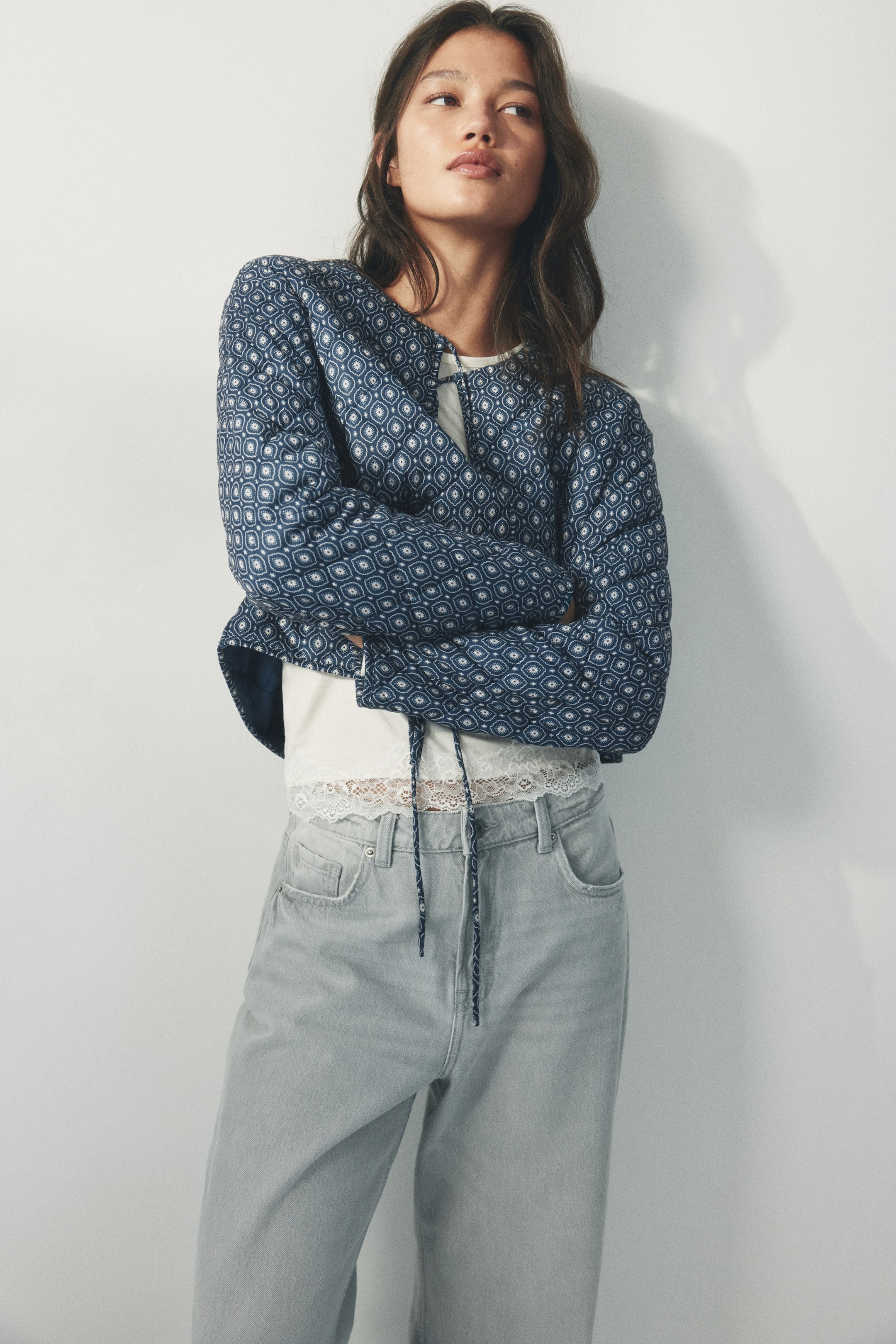 View larger image: A quilted jacket, featuring a navy blue fabric with a white geometric pattern, is displayed with a cropped length, a round neckline, and a drawstring hem. Its long sleeves are visible as it is being worn over a white top.