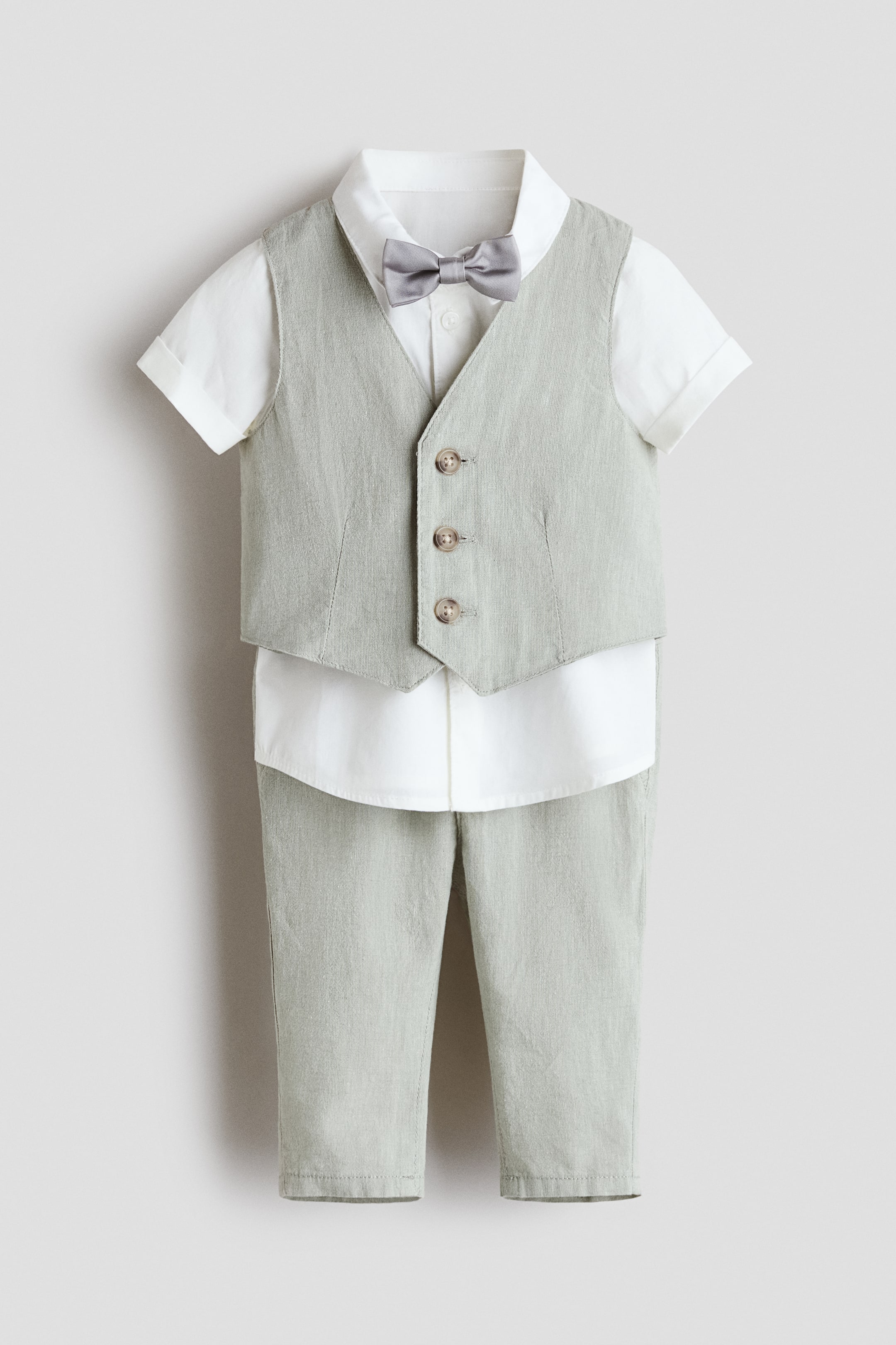 View larger image: A smart ensemble is being presented from the front, featuring a light green waistcoat and matching trousers in a soft weave. The waistcoat is shown with three buttons down the front, worn over a white short-sleeved shirt with a turn-down collar and sewn-in turn-ups at the cuffs. A grey pre-tied bow tie completes the outfit.