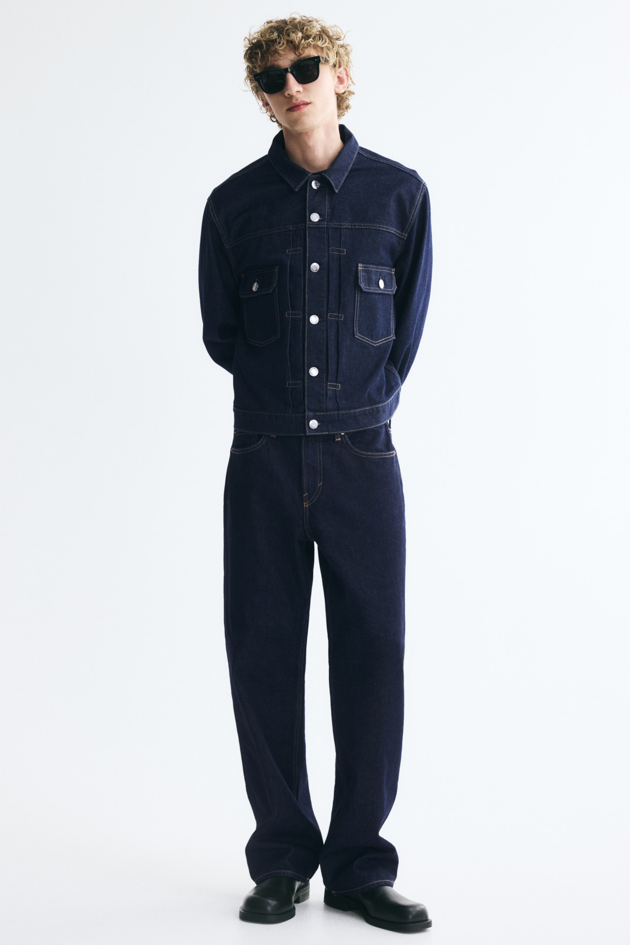 View larger image: Dark blue denim jeans with a relaxed, straight-leg fit are worn by the model. The jeans are styled with a matching buttoned-up dark denim jacket, black sunglasses, and black shoes.