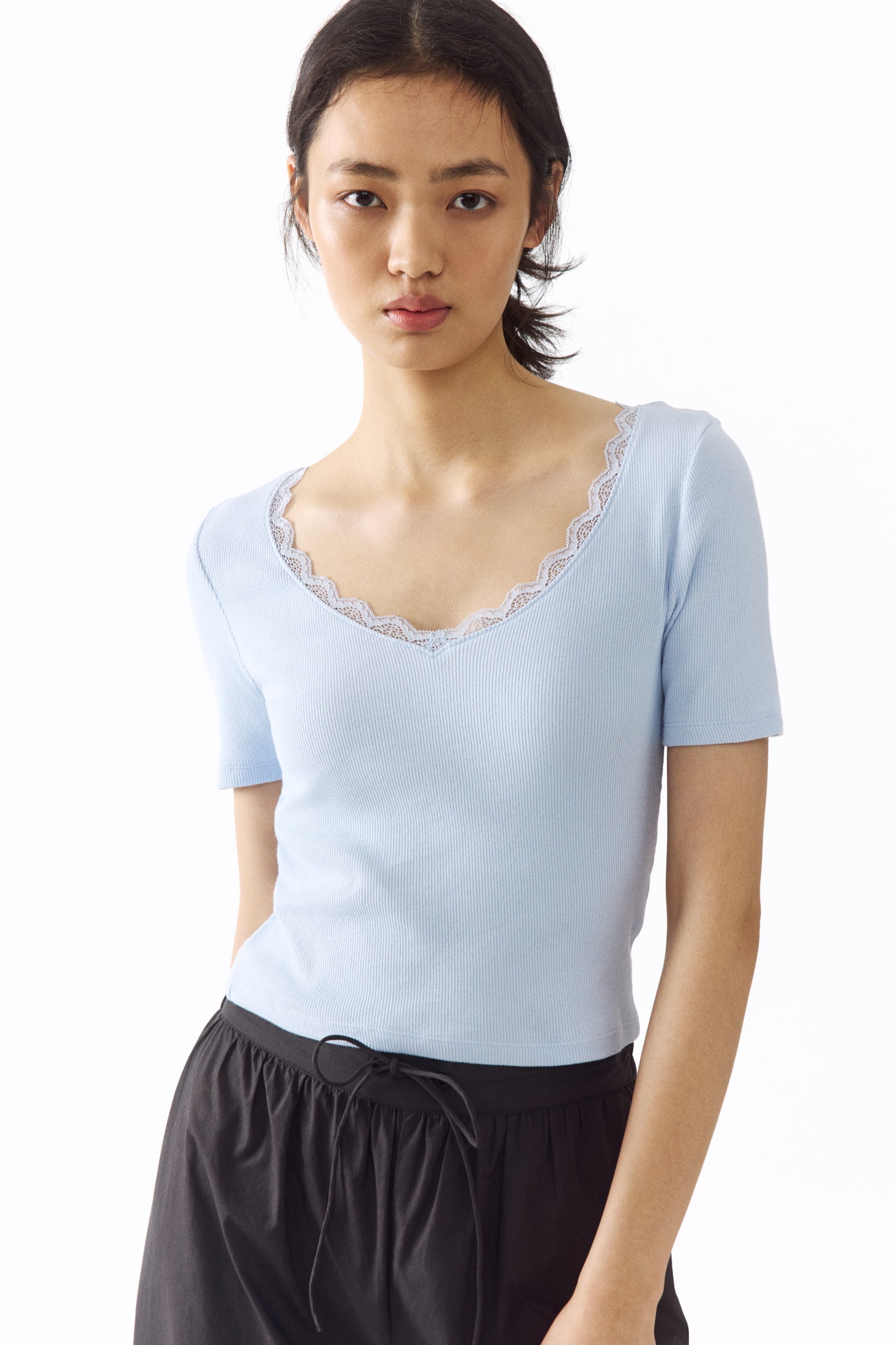 הצגת תמונה גדולה יותר: חולצה צמודה עם שרוולים קצרים בצבע כחול בהיר, בסריגת ריב עדינה, עם מחשוף V רחב ועמוק עם עיטור תחרה, נלבשת על ידי הדוגמנית.