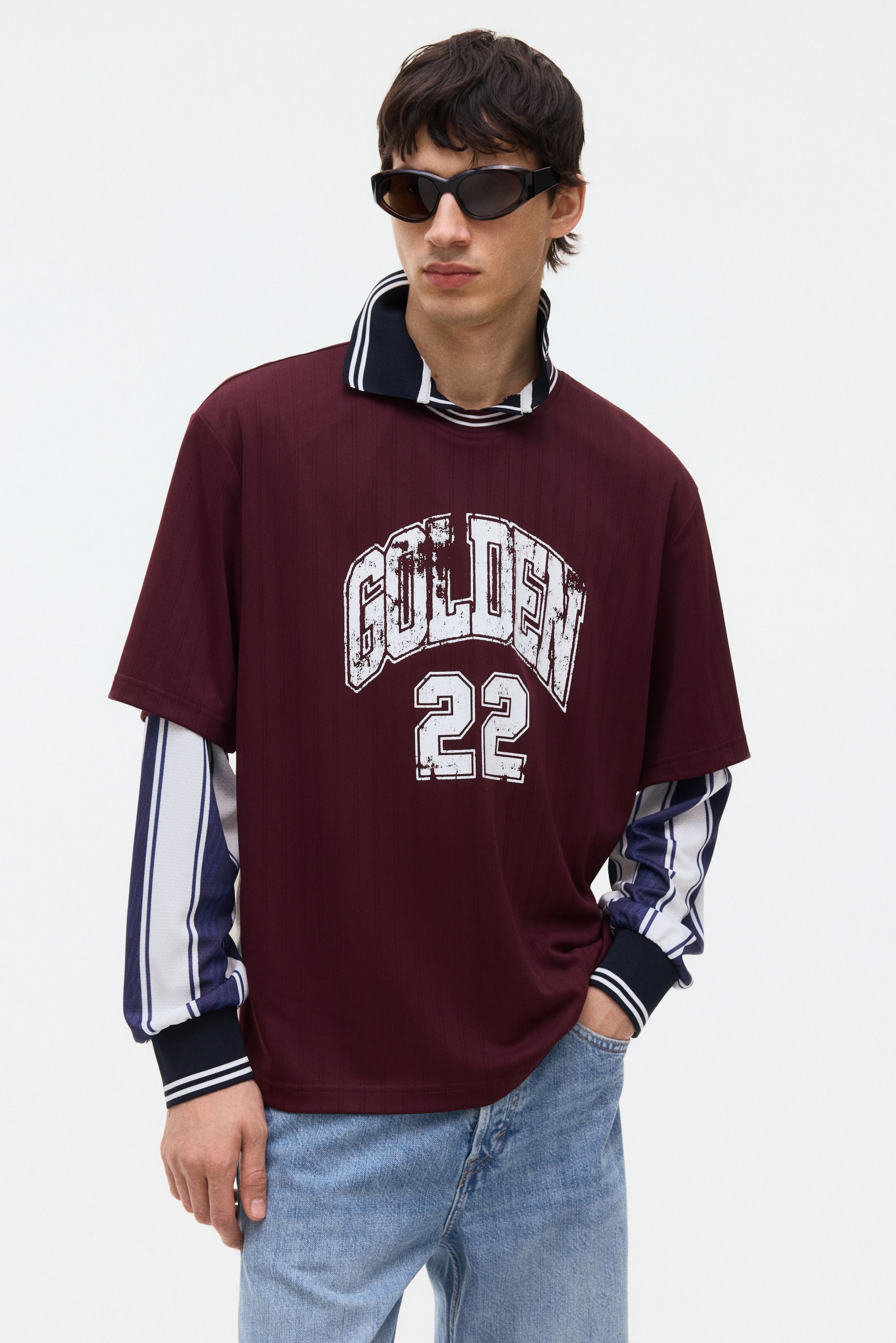 Visa större bild: En vinröd tröja i jersey-stil med fina vertikala ränder och ett slitet vitt "GOLDEN 22"-tryck presenteras framifrån, något vinklad åt höger. Den har en stående marinblå polokrage med vita ränder och är lagrad över en marinblå och vit randig långärmad skjorta med marinblå ribbade muddar.