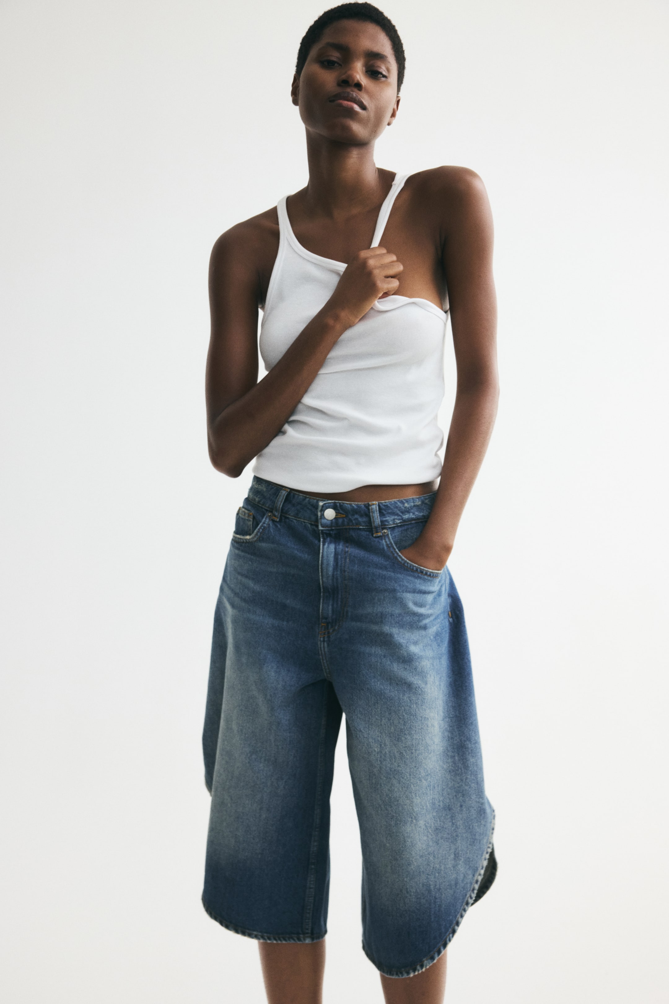 Größeres Bild anzeigen: 5-Pocket-Shorts aus festem Baumwoll-Denim, mit abgenutzten Details, einer normalen Taille mit sichtbarem Knopfverschluss, überlappenden Seiten und abgerundeten Säumen, werden von dem Model getragen, das eine Hand in eine Vordertasche steckt.
