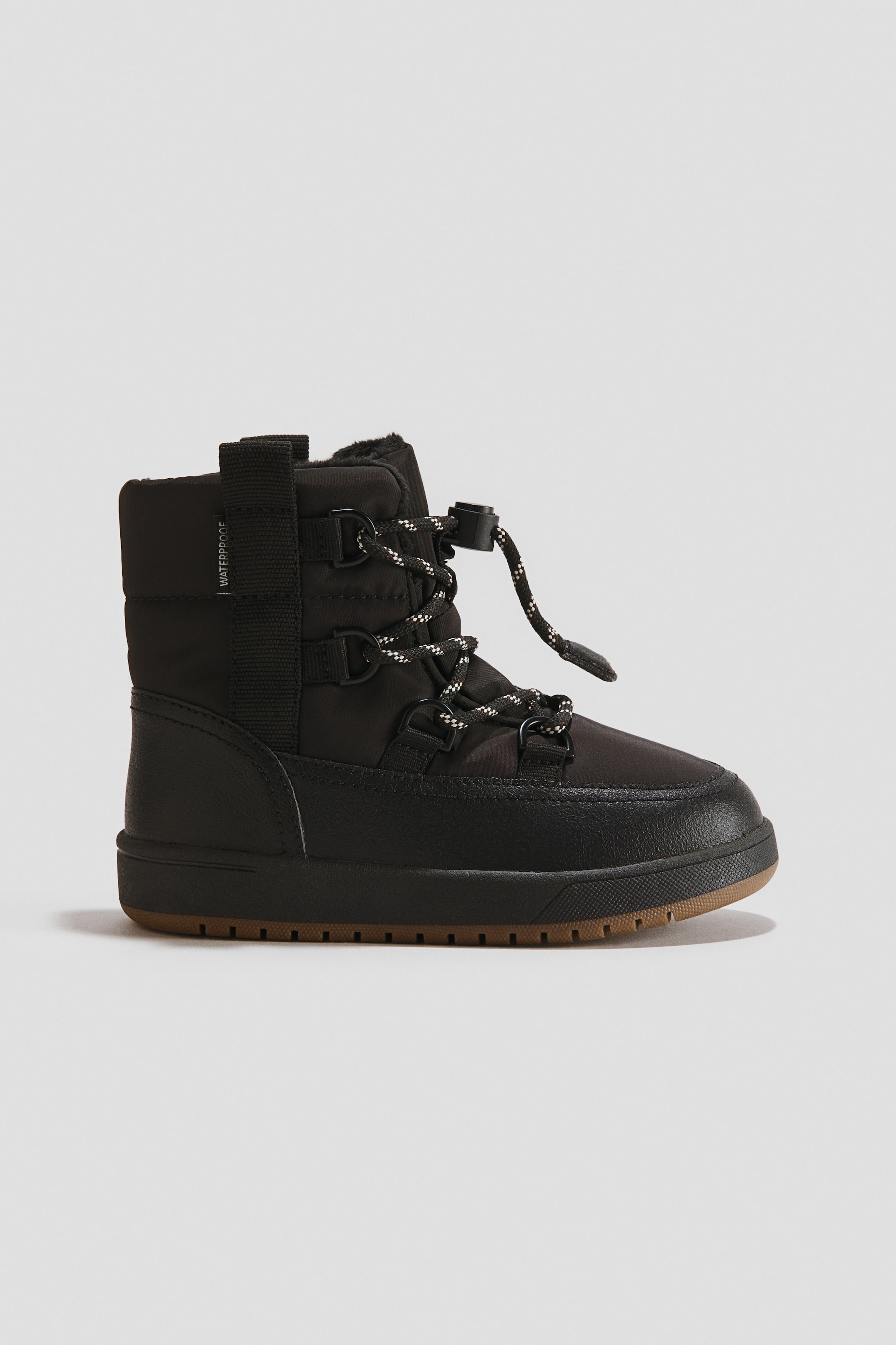 View larger image: A black warm-lined waterproof snow boot is displayed with its padded upper and visible furry lining at the opening. It features a 'waterproof' label on the side, a lace-up front with D-rings, black and white patterned laces, and a toggle fastening, resting on a thick black sole with visible brown tread.