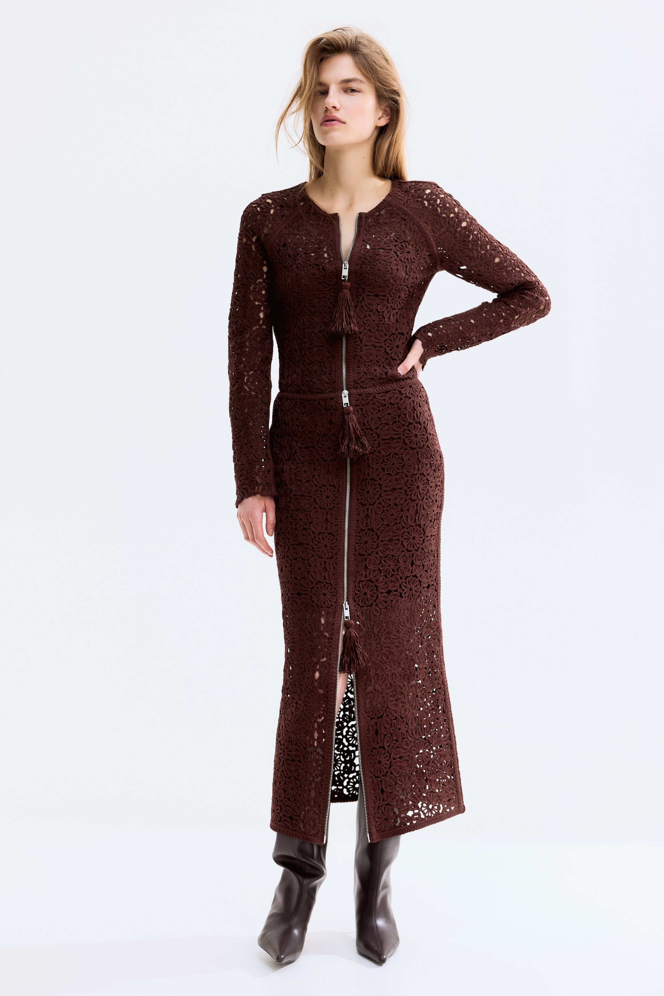 Ver imagen más grande: Un vestido maxi de encaje marrón presenta un patrón floral calado, mangas largas y un escote redondo. Un cierre frontal de cremallera de longitud completa con dos tiradores de borlas recorre el centro, y una prenda negra es visible debajo del encaje en la parte inferior.