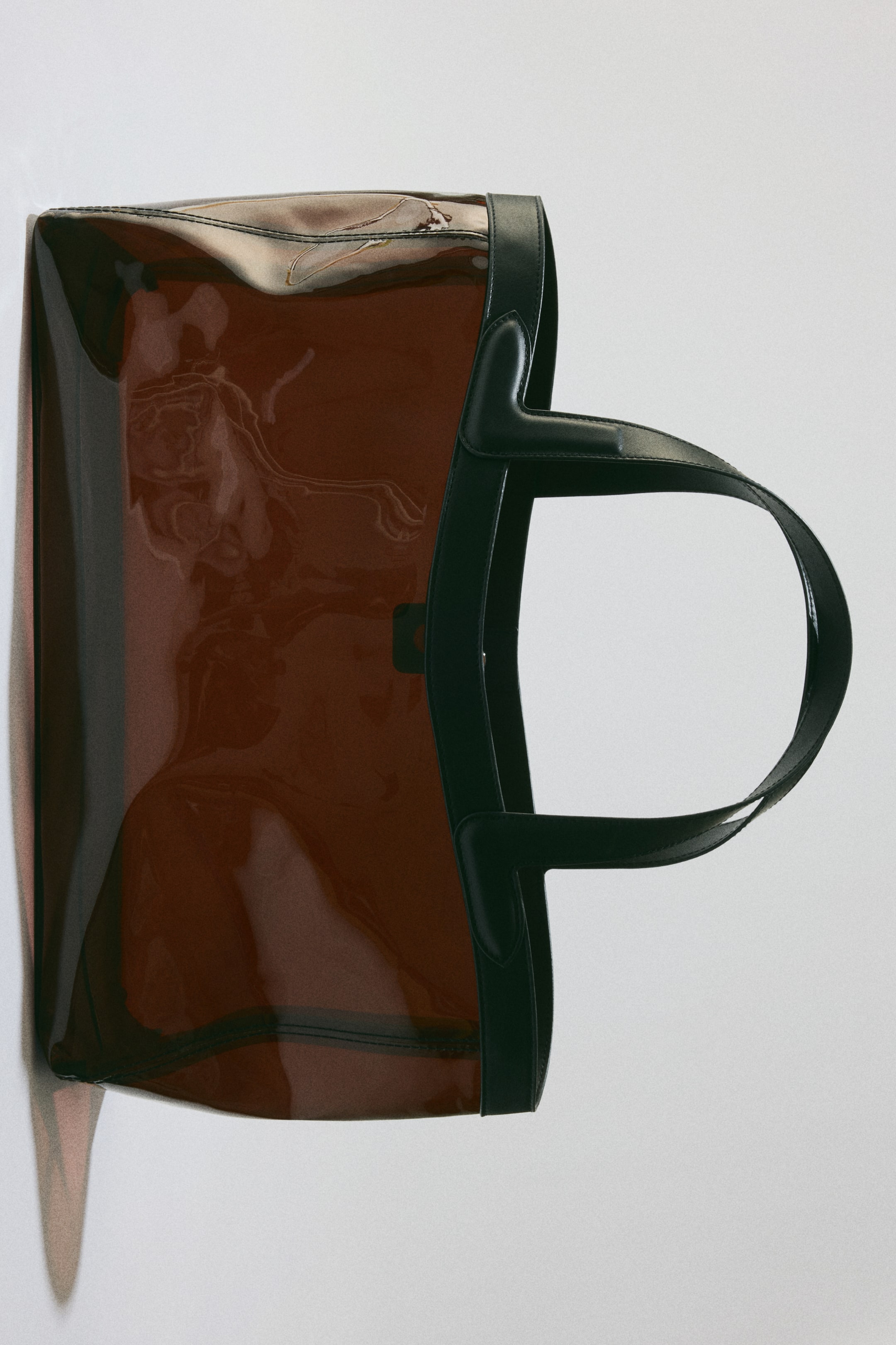 View larger image: This tote bag showcases a translucent reddish-brown body complemented by black trim that outlines the top edge and forms two structured handles. The zipper closure is prominently visible along the top opening, with the bag positioned horizontally and its opening oriented to the right.