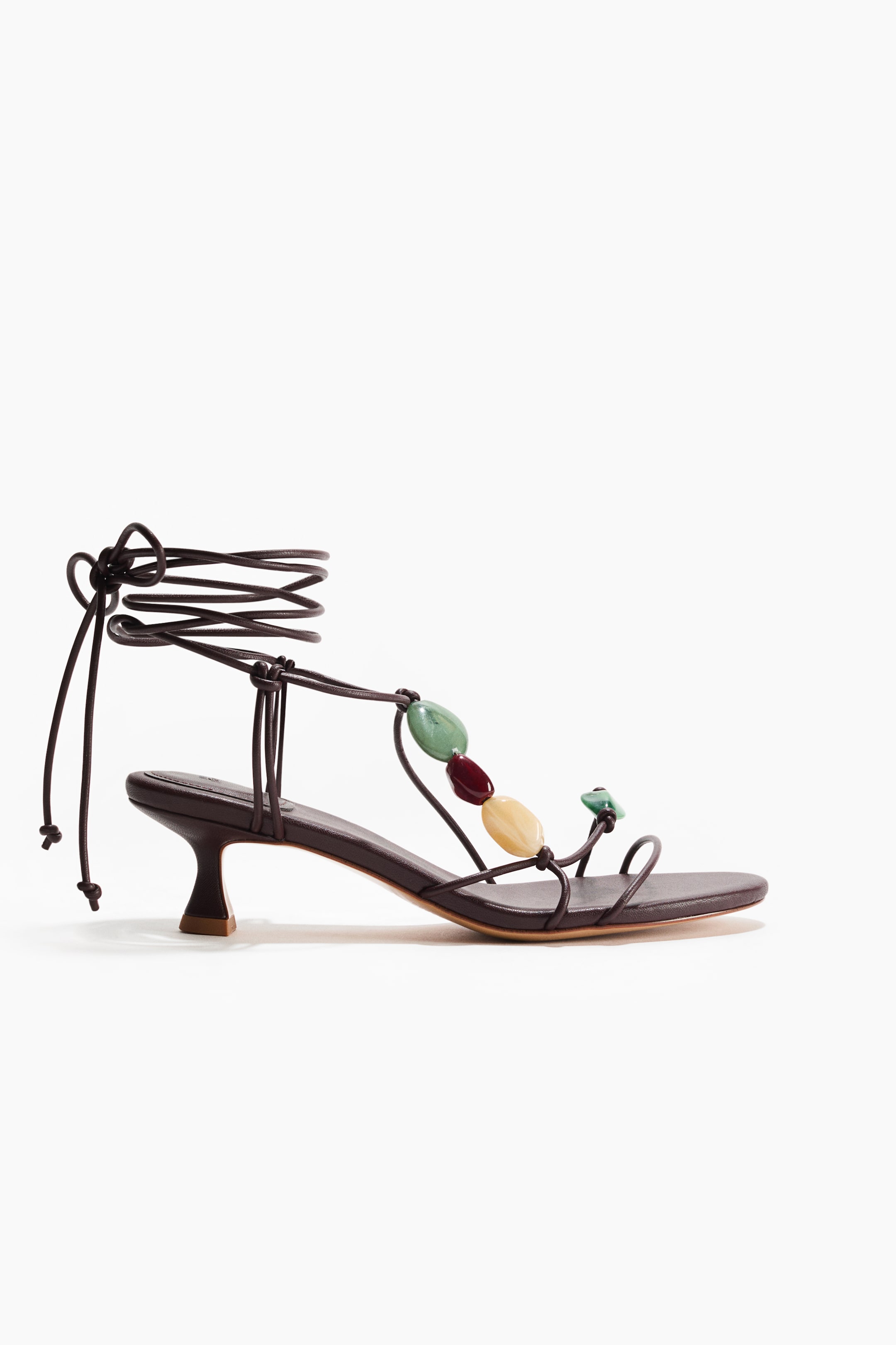 View larger image: A sandal in dark brown coated fabric features knotted spaghetti foot straps adorned with colorful plastic beads. Long spaghetti ties wrap around the ankle, and the sandal showcases an almond toe and a flared kitten heel.