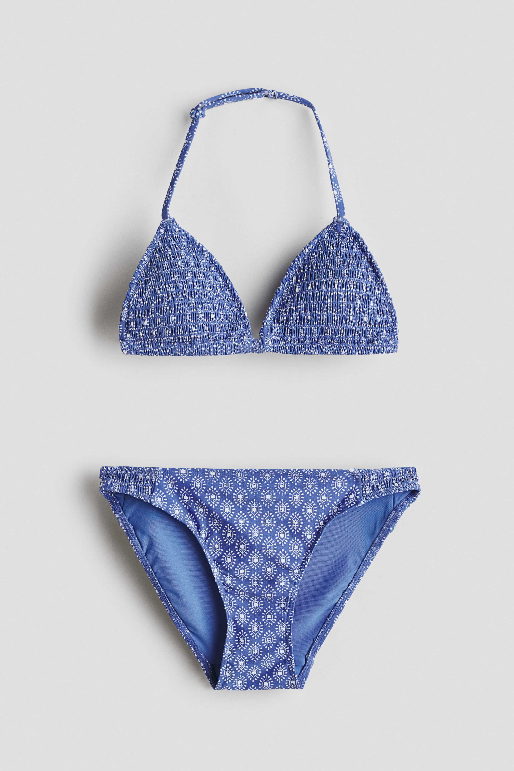 Grösseres Bild ansehen: Ein Bikini-Set wird von VORNE gezeigt, mit einem blauen Halter-Triangle-Top mit gesmokten Cups. Die passenden Bikini-Slips präsentieren ein weißes geometrisches Muster auf Blau, mit gerafften Seiten und einem festen blauen Futter.