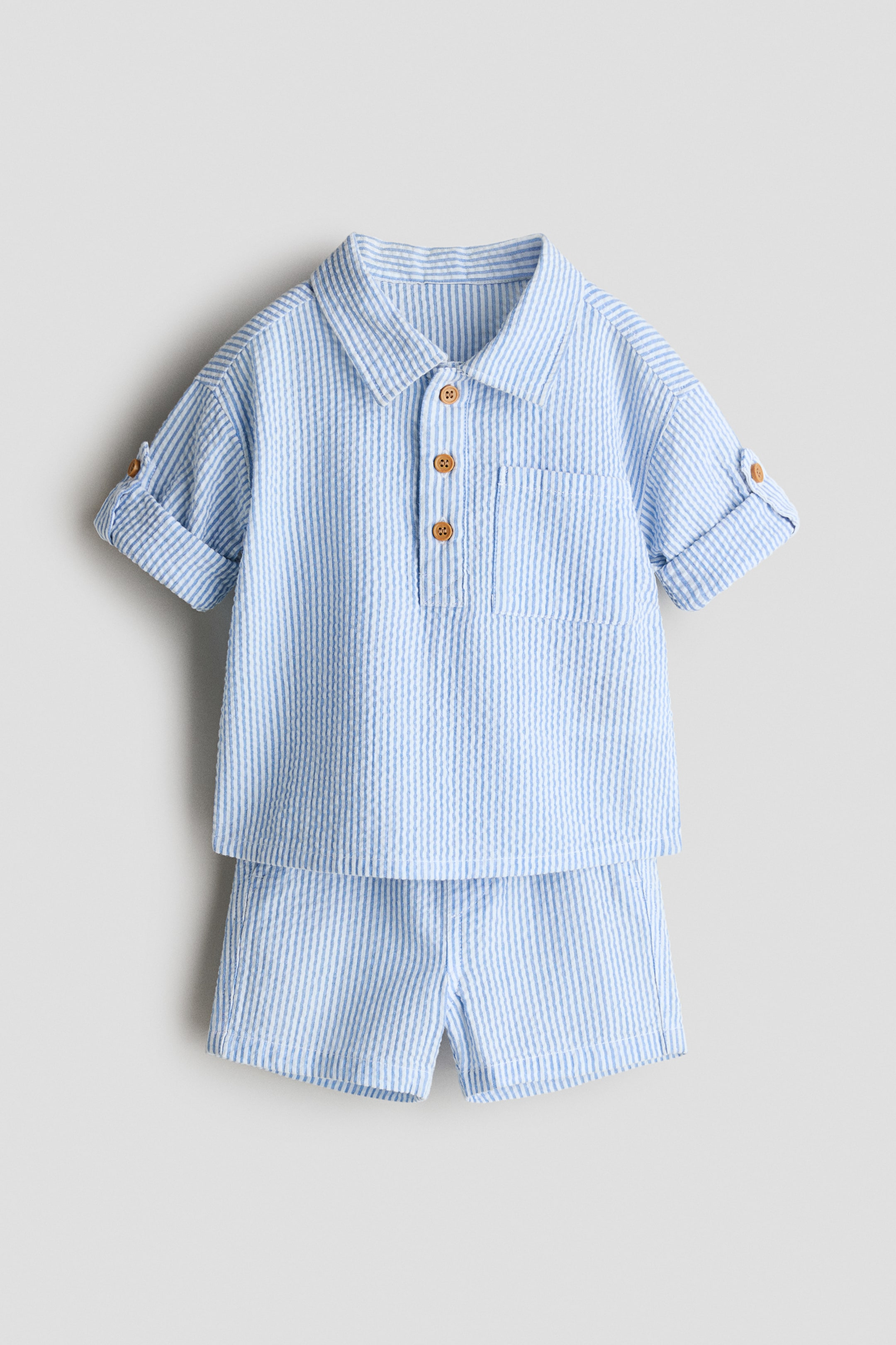 Agrandir l'image: La tenue en seersucker à rayures bleu clair et blanc pour bébé est présentée de face, comprenant une chemise à col et un short assorti. La chemise est conçue avec une patte de boutonnage à trois boutons en bois, une poche poitrine unique, et des manches courtes retroussées et fixées avec une patte boutonnée.
