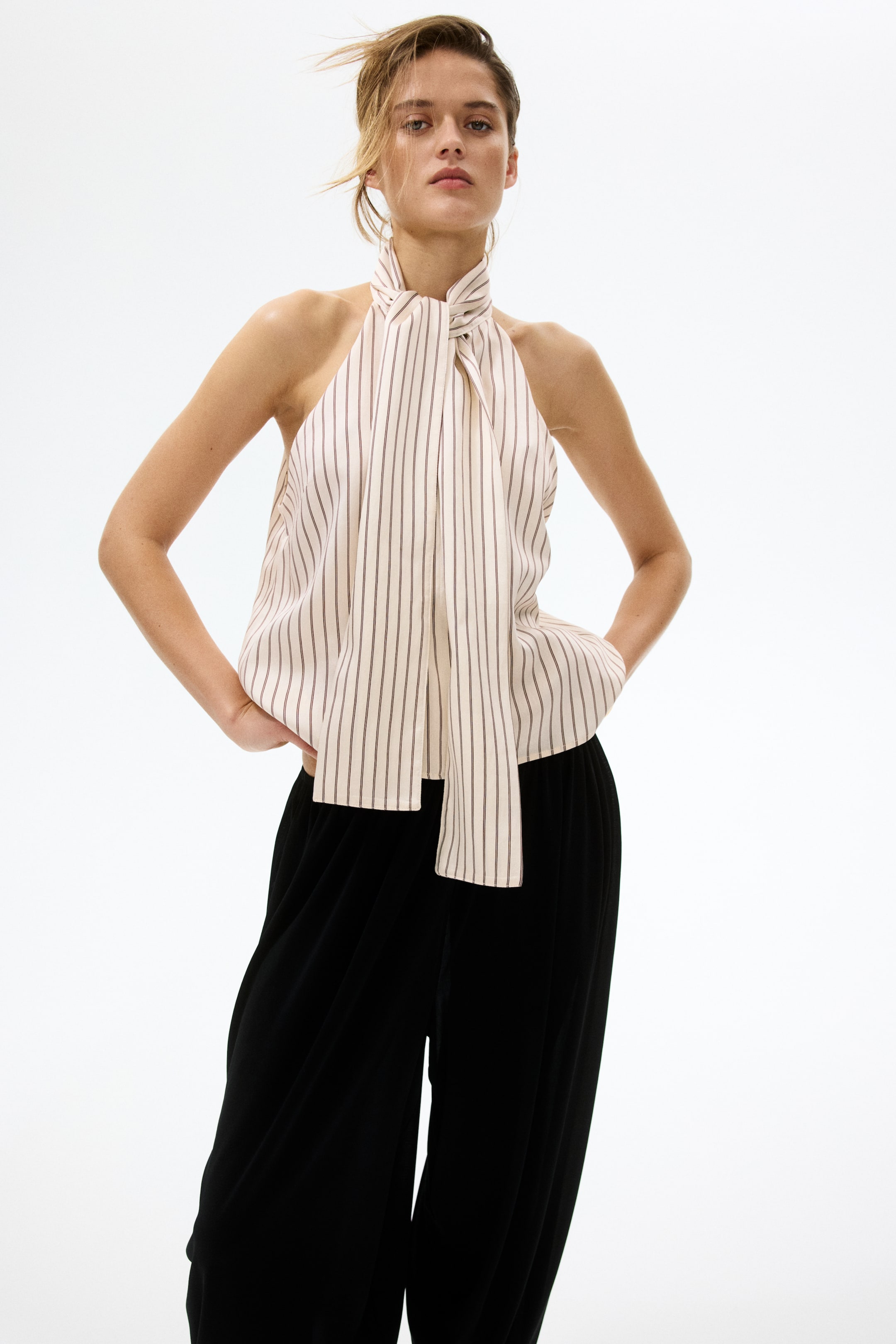 View larger image: The model is wearing a sleeveless striped top with a bow collar tied at the neck, creating a V-shaped front opening. The top is narrowly cut at the top and paired with black wide-leg pants.