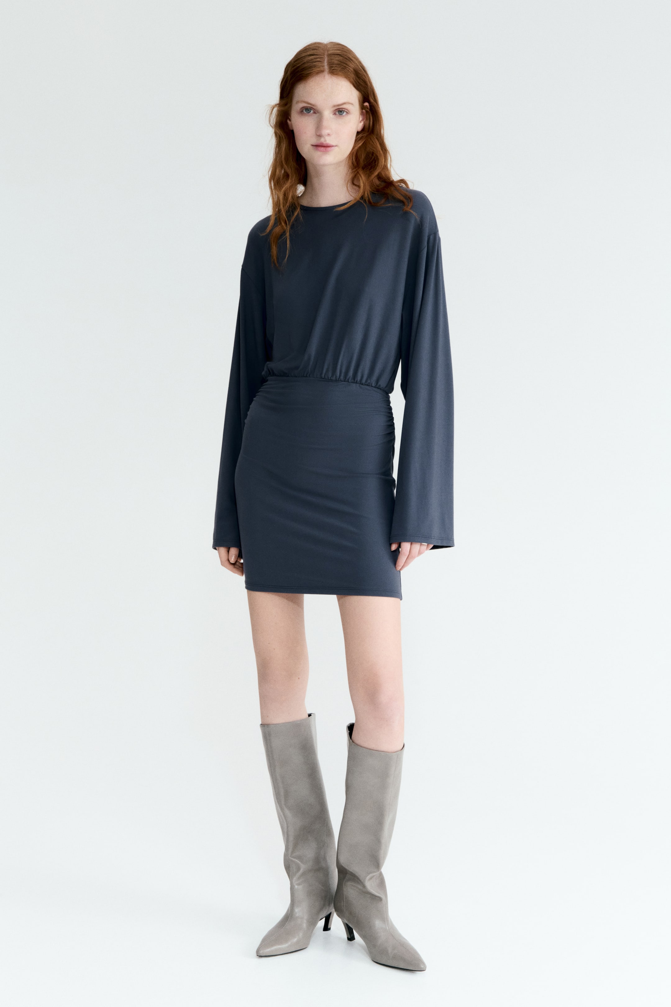 View larger image: A dark gray-blue mini dress is showcased front-on, highlighting a loose-fitting top with long, relaxed sleeves and a crew neck. The dress cinches at the waist with gathering, and its fitted mini skirt is ruched on the sides.