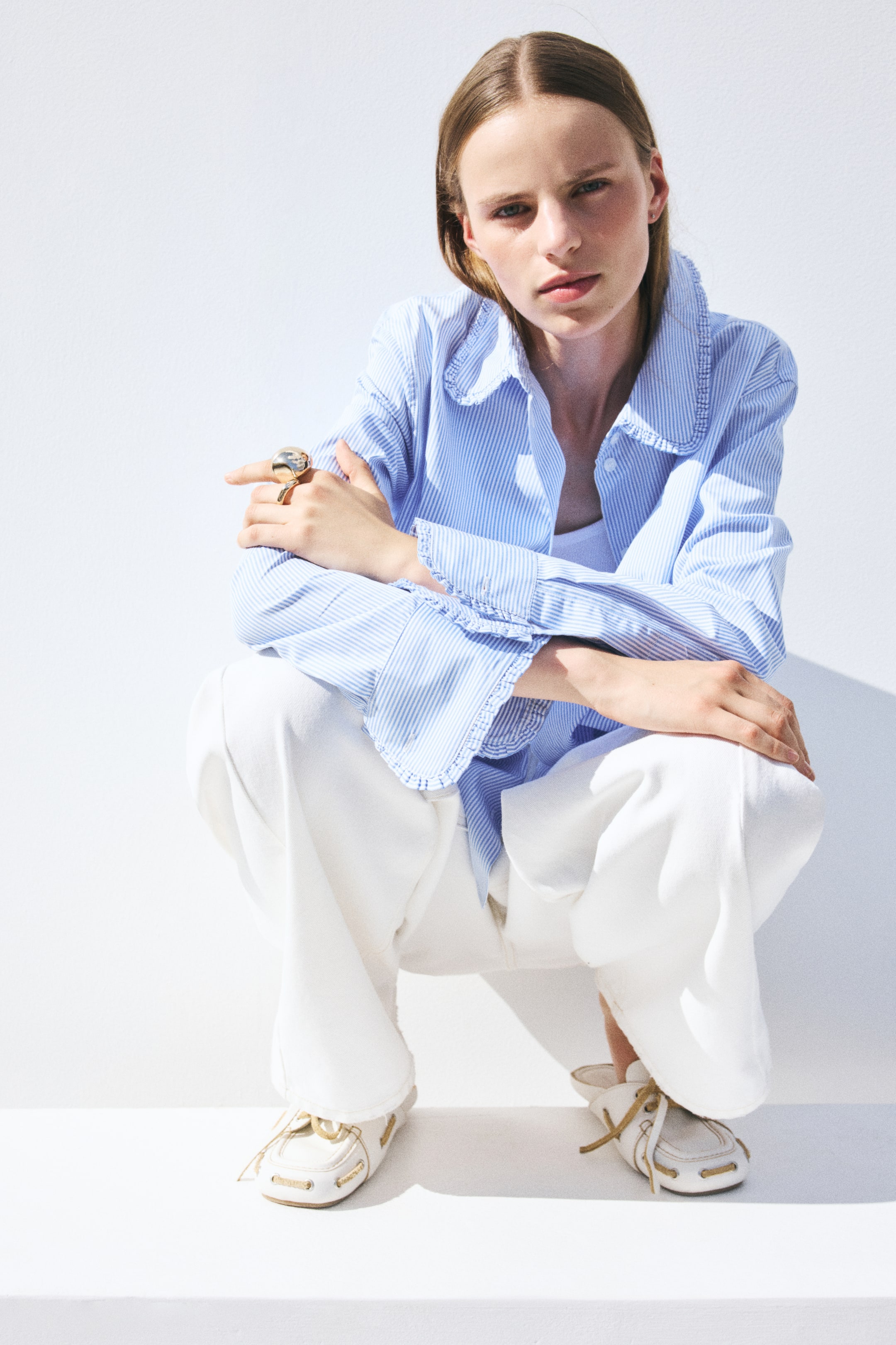 View larger image: A blue and white striped long-sleeve shirt with a frill-trimmed collar and cuffs is worn open by the model, who is crouching. The shirt is layered over a white top and paired with white trousers.