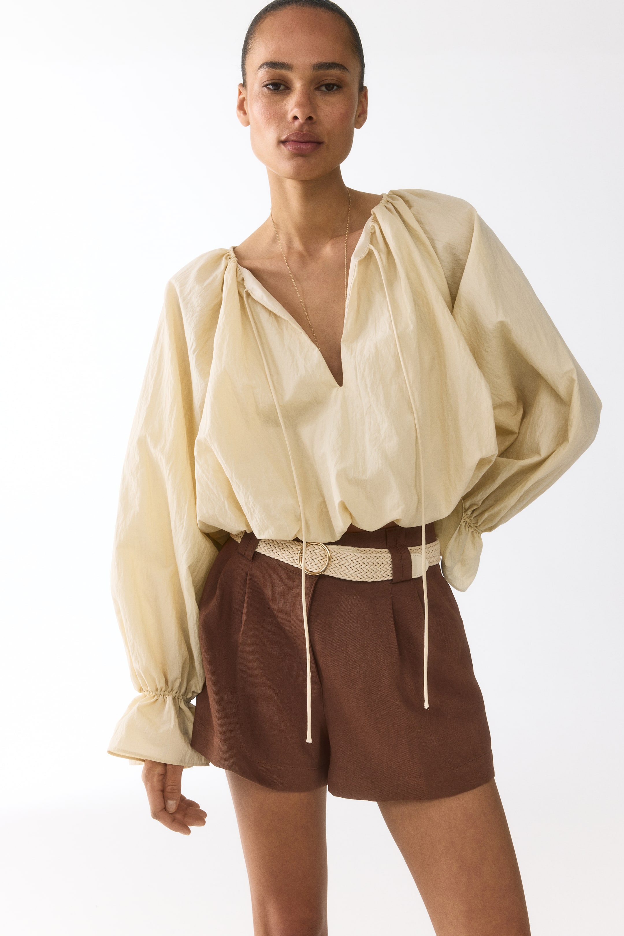 Visa större bild: En ljus beige voluminös blus visar upp en V-ringning med knytband och rynkade, fullängdsärmar som avslutas med volangmanschetter. Den är stylad med bruna plisserade shorts, som hålls uppe av ett vävt ljusfärgat bälte med ett cirkulärt spänne.