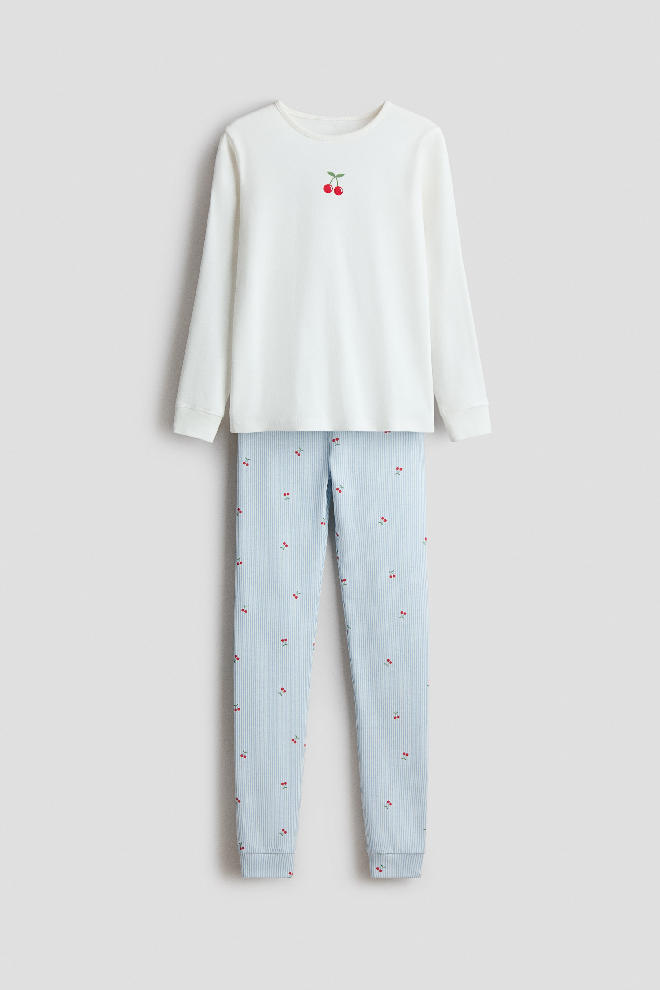 View larger image: A two-piece pajama set is presenting front, featuring a white long-sleeved top with a crew neck and a small red cherry embroidery on the front chest. The coordinating light blue pants feature thin white vertical stripes and scattered red cherry motifs, and both the top and pants finish with ribbed cuffs.