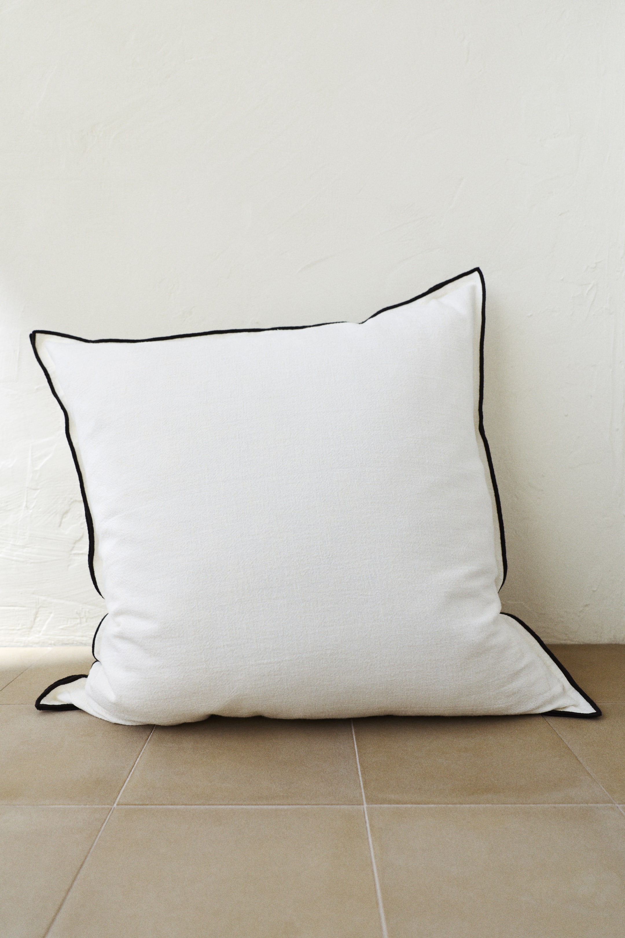 Ver imagem maior: A almofada branca apresenta um debrum preto ao longo de todas as suas bordas, exibindo um tecido suave e ligeiramente texturizado. Está inclinada ligeiramente, com a frente voltada para a direita.