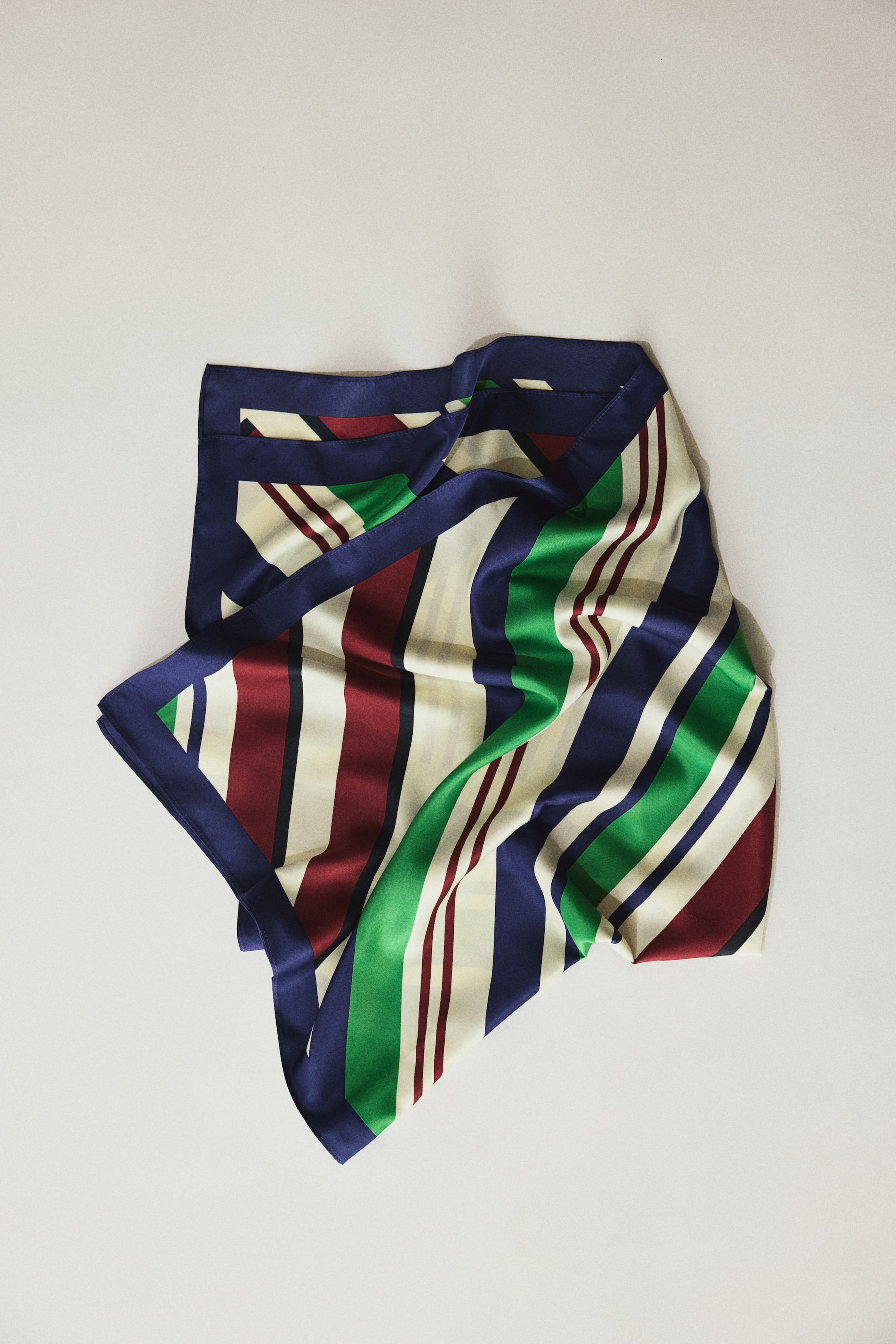View larger image: A square scarf features a striped pattern in navy blue, cream, dark red, and green. It has a solid navy-blue border and is shown folded to showcase its smooth, reflective fabric.