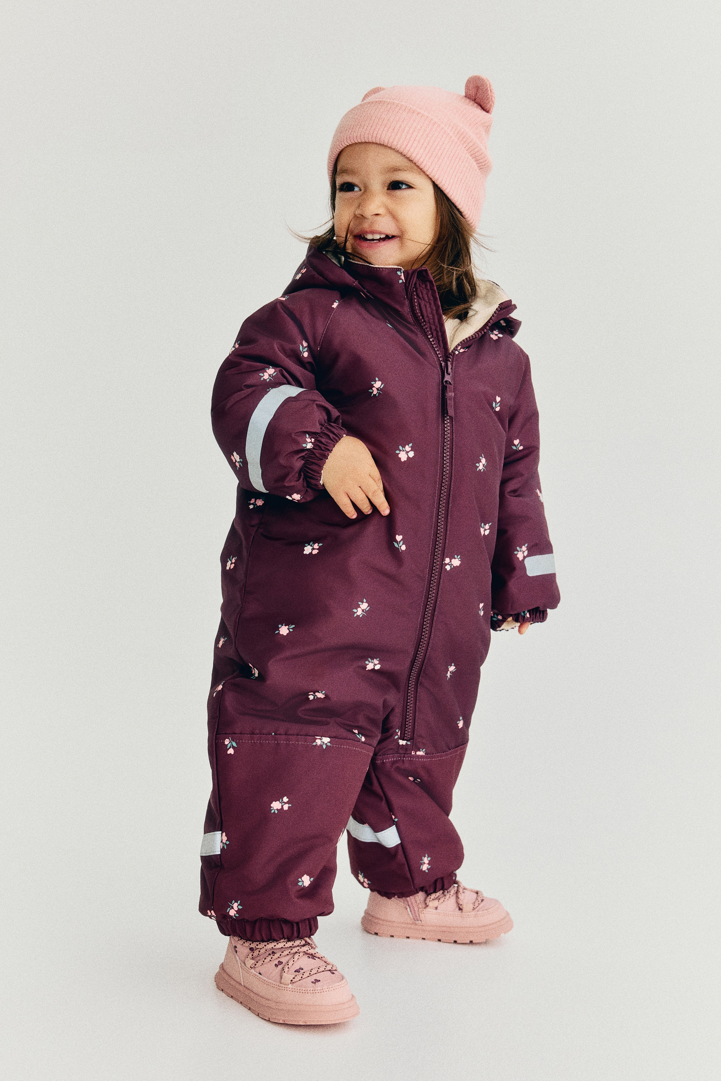 A dark red padded all-in-one suit, patterned with small pink and white flowers, is facing forward, featuring a cream fleece-lined detachable hood and a front zipper closure. Reflective strips are visible on the sleeves and lower legs, along with elasticated cuffs and hems, reinforced sections on the knees and lower back, and detachable stirrup straps extending from the leg openings.