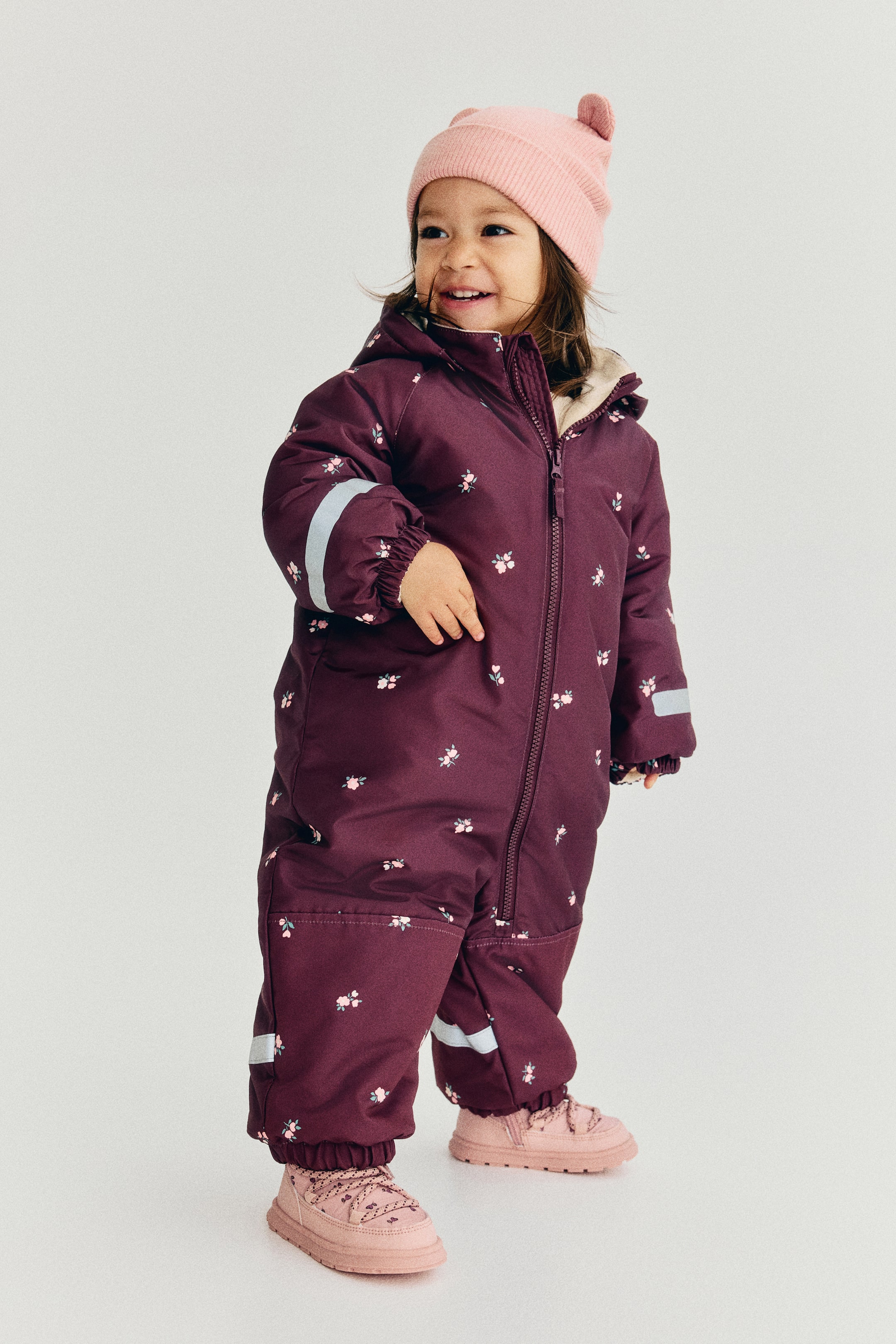 View larger image: A burgundy one-piece snowsuit featuring a small pink floral pattern is shown from the front, highlighting a full-length zipper and an attached hood lined with cream-coloured fleece. The garment features elasticated cuffs and ankles with reflective silver safety bands.