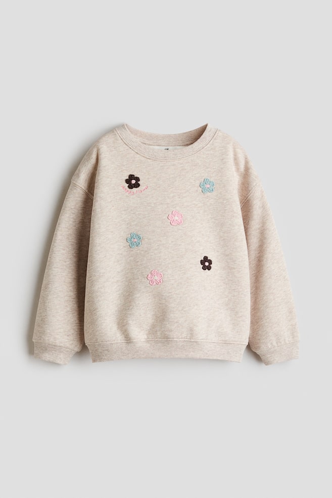 Se muestra una sudadera melange beige claro vista de frente, con cuello redondo acanalado, hombros caÃ­dos y puÃ±os y basta acanalados. La parte frontal presenta pequeÃ±os detalles de flores bordadas dispersas en rosa, azul claro y cafÃ© oscuro. - 1