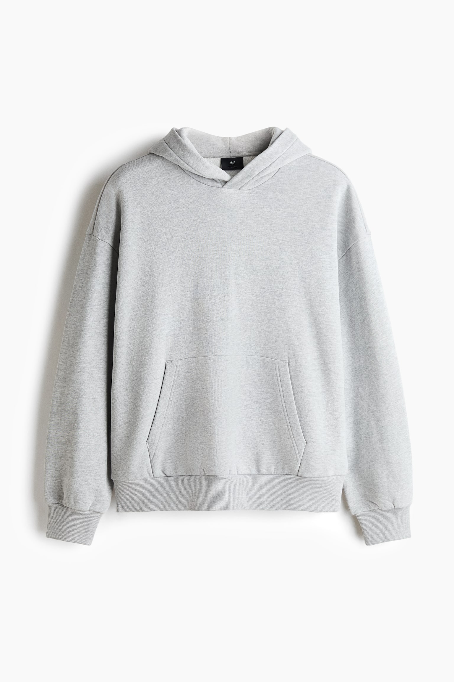 Een oversized lichtgrijze hoodie hangt recht van voren in beeld en toont een omslagkap, verlaagde schoudernaden en lange mouwen met geribde boorden. Het kledingstuk heeft een ruime kangaroozak aan de voorkant en een geribde zoom, wat het losse, relaxte silhouet benadrukt.