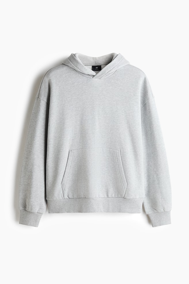 Ein oversized hellgrauer Hoodie hÃ¤ngt in frontaler Ansicht und zeigt eine Ã¼berlappende Kapuze, Ã¼berschnittene Schultern sowie lange Ãrmel mit RippbÃ¼ndchen. Das KleidungsstÃ¼ck verfÃ¼gt Ã¼ber eine groÃzÃ¼gige KÃ¤ngurutasche vorne und einen gerippten Saum, wodurch seine lockere, entspannte Silhouette betont wird. - 1