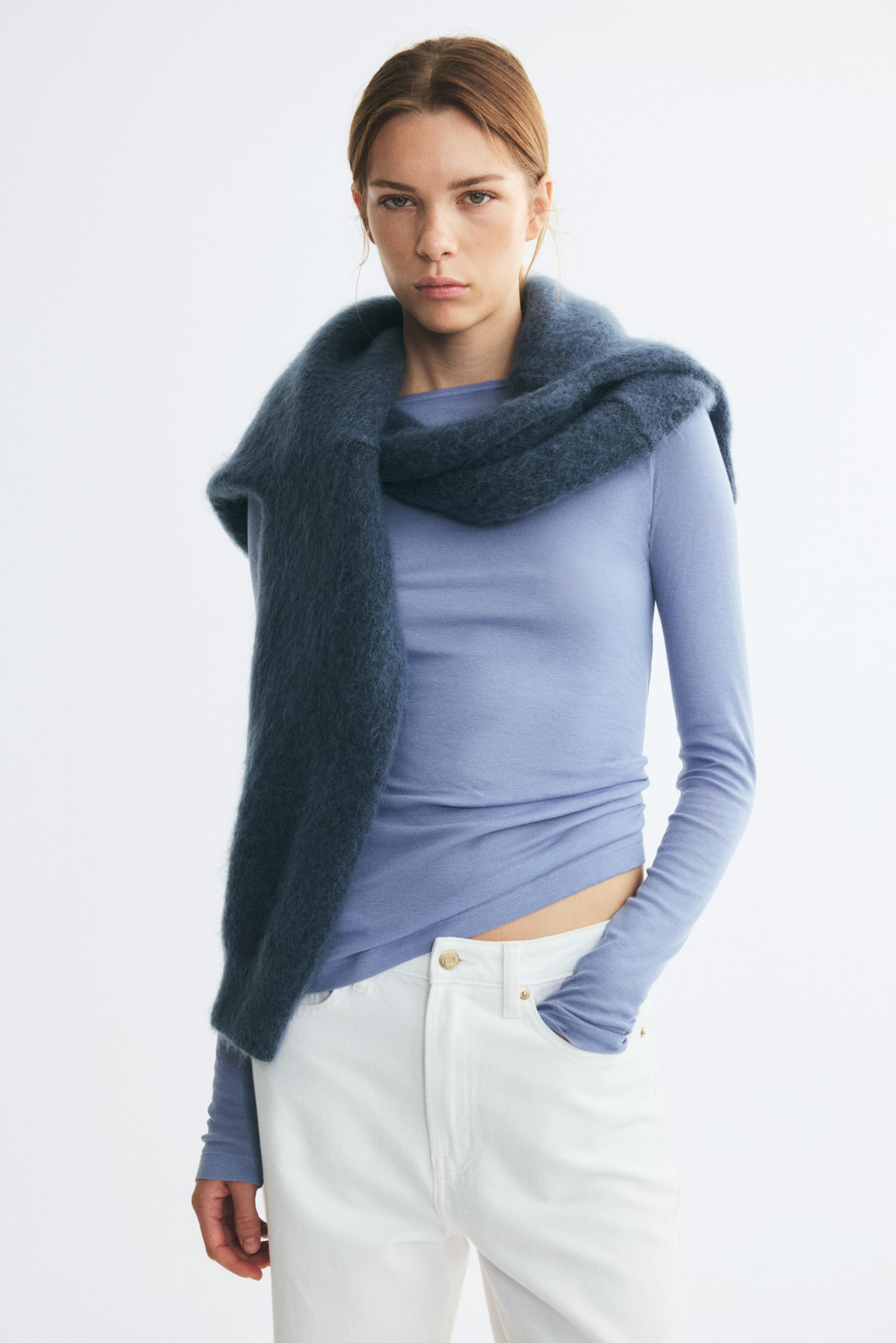 View larger image: The model is wearing a light blue long-sleeved top, styled with a textured dark blue knit sweater draped over the shoulders. The top is paired with white high-waisted jeans, with one hand resting in a front pocket.