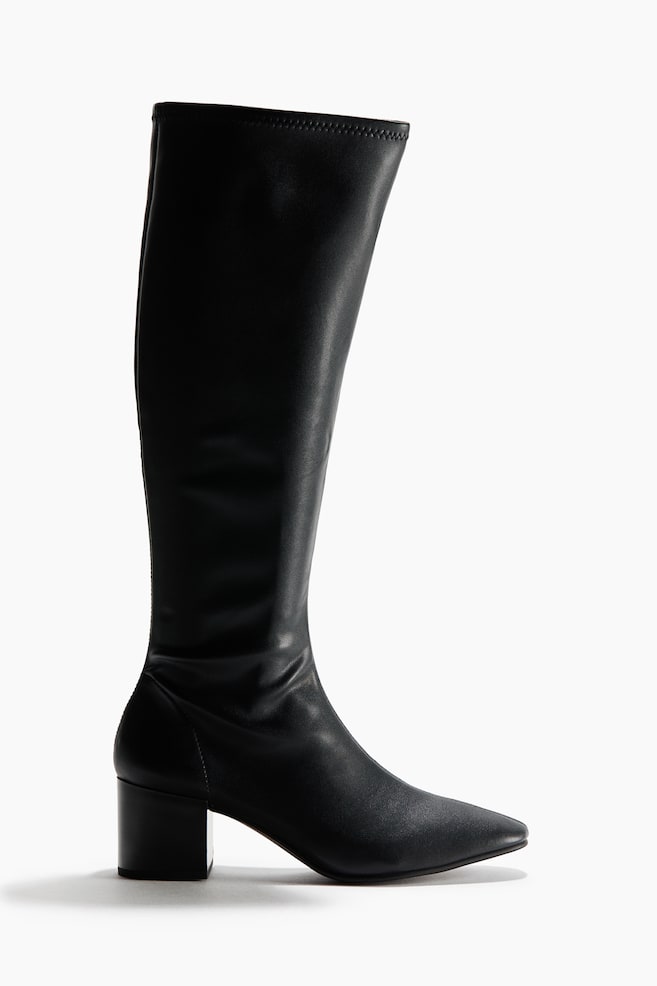 A black knee-high boot is standing in a side profile view facing right, showcasing a pointed toe and a low block heel. The boot is featuring a fitted, smooth shaft that extends up to the knee with a clean, minimalist design. - 1