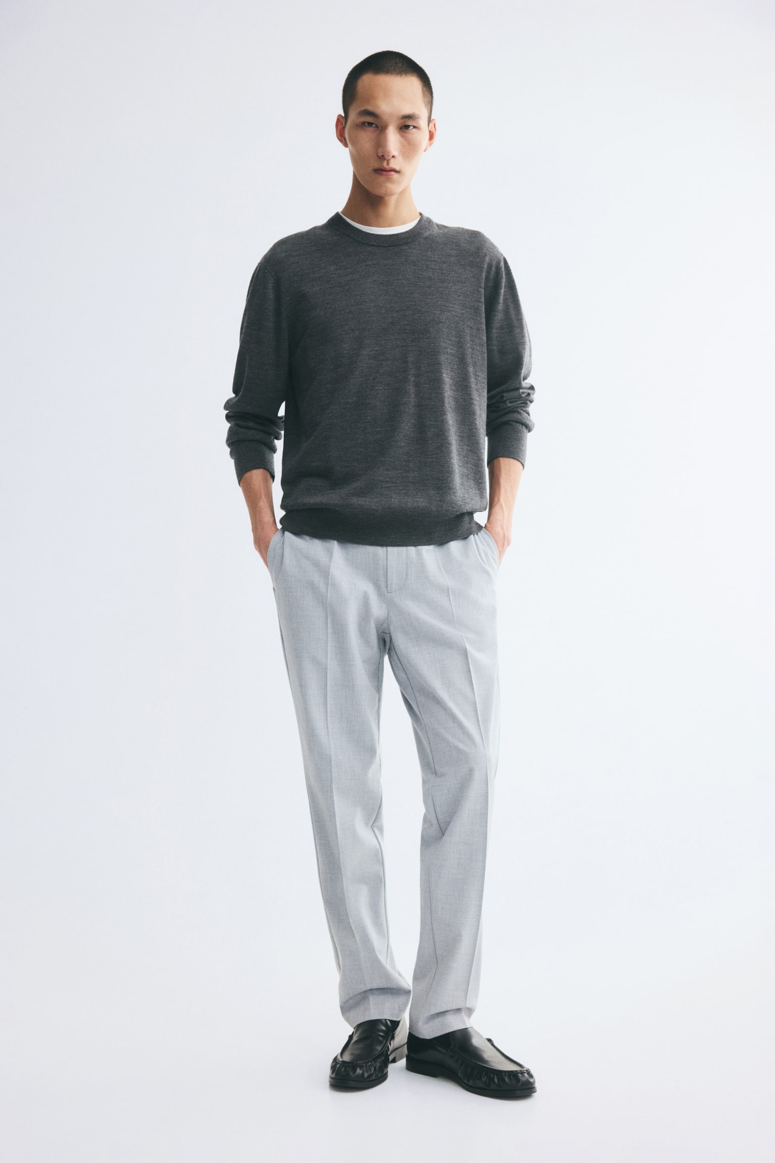Tailored light grey joggers in a woven fabric are being worn by the model, featuring a slim fit and sharp creases running down the front of the legs. The model is standing with hands resting in the side pockets, styling the trousers with a dark grey crewneck sweater and black leather loafers. - 1