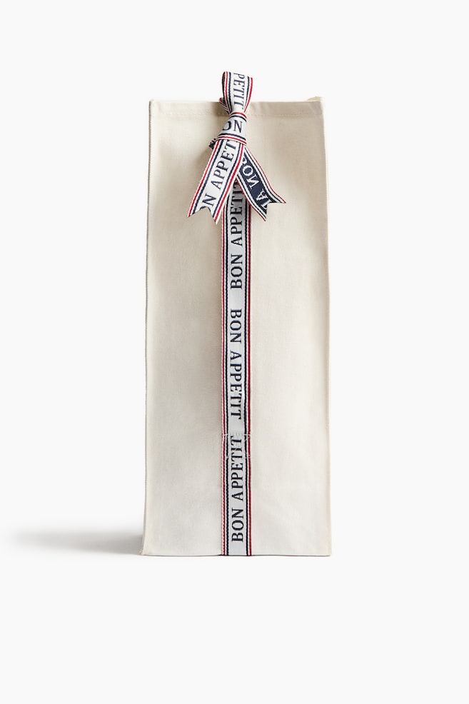 A cream fabric bottle bag stands upright, facing forward. It features a red, white, and blue striped ribbon tied in a bow at the top, with the ribbon extending vertically down the front and reading "BON APPÉTIT" repeatedly. - 1