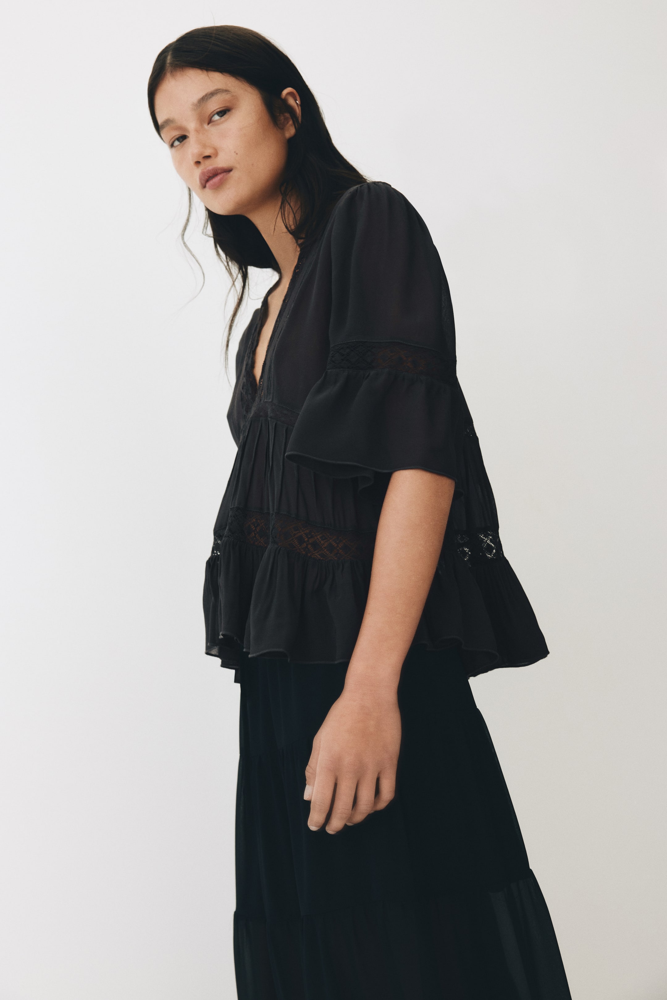 View larger image: The model is wearing a black sheer chiffon blouse, showcasing a lace-trimmed V-neckline, lace insets on the sleeves and bodice, pintucks under the bust, and 3/4-length sleeves with flared cuffs, flowing to a gently flared hem.