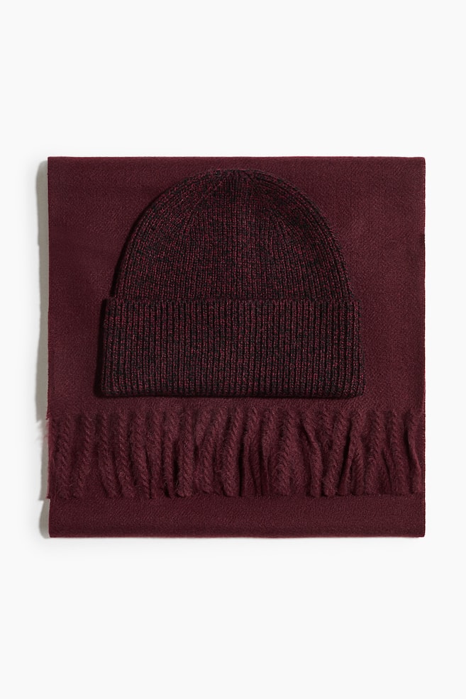 Se muestra de frente un conjunto de accesorios de invierno en color burdeos, con un gorro de punto jaspeado con puño doblado y acanalado, apoyado sobre una bufanda burdeos doblada con ribete de flecos retorcidos. - 1