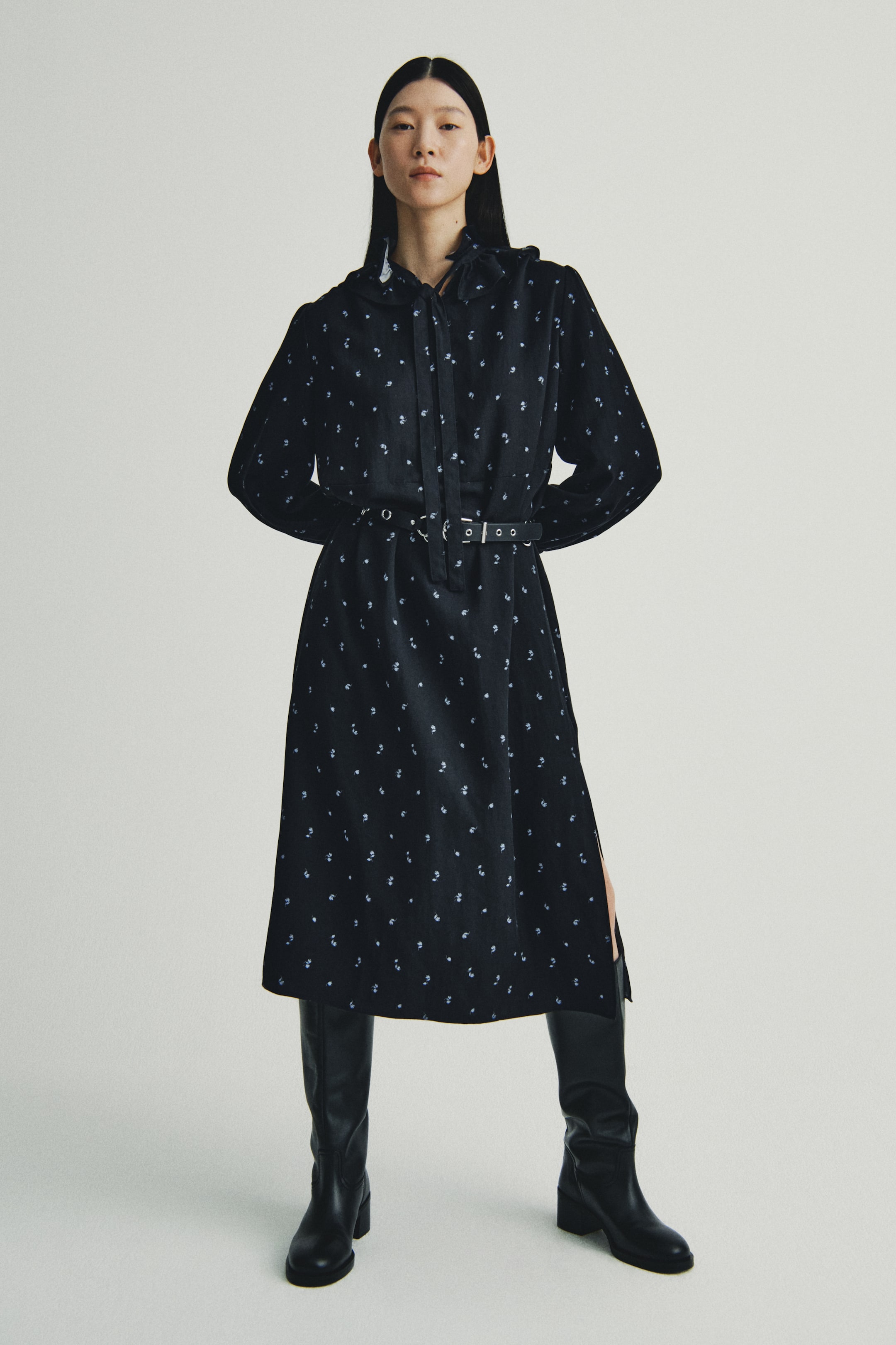 Xem ảnh lớn hơn: Một chiếc váy midi màu đen phía trước có họa tiết trừu tượng nhỏ, màu sáng. Nó được thiết kế với tay áo dài, cổ nhọn, và chi tiết thắt nơ ở cổ, ôm eo bằng một chiếc thắt lưng màu đen với khóa bạc. Phần váy có độ dài midi với một đường xẻ bên trái.