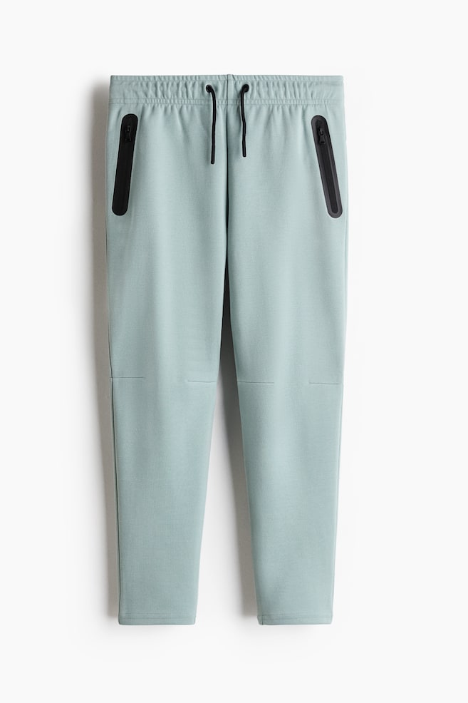 Los pantalones de chándal en color azul verdoso claro se muestran en vista frontal, con una cintura elástica que incluye un cordón negro y dos bolsillos con cremallera en ángulo de color negro. Se pueden apreciar detalles de costuras horizontales justo debajo de las rodillas en ambas piernas. - 1