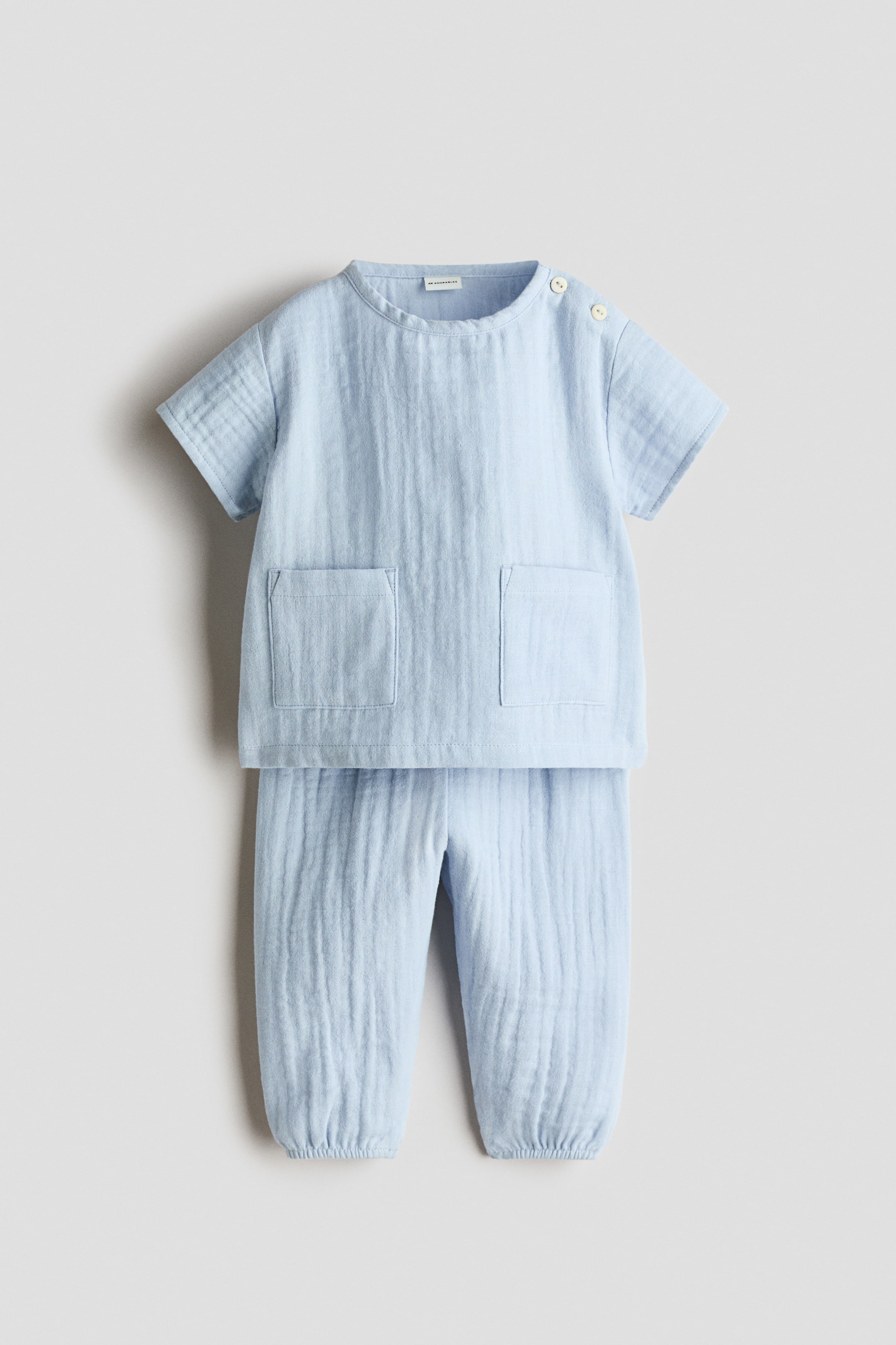 Ampliar la imagen: Un conjunto de dos piezas en azul claro, hecho de tela arrugada, se muestra en vista frontal. La blusa de manga corta presenta un escote redondo, dos botones de color claro en el hombro izquierdo y dos bolsillos de parche al frente, mientras que los pantalones muestran una cintura elástica y dobladillos elásticos fruncidos.
