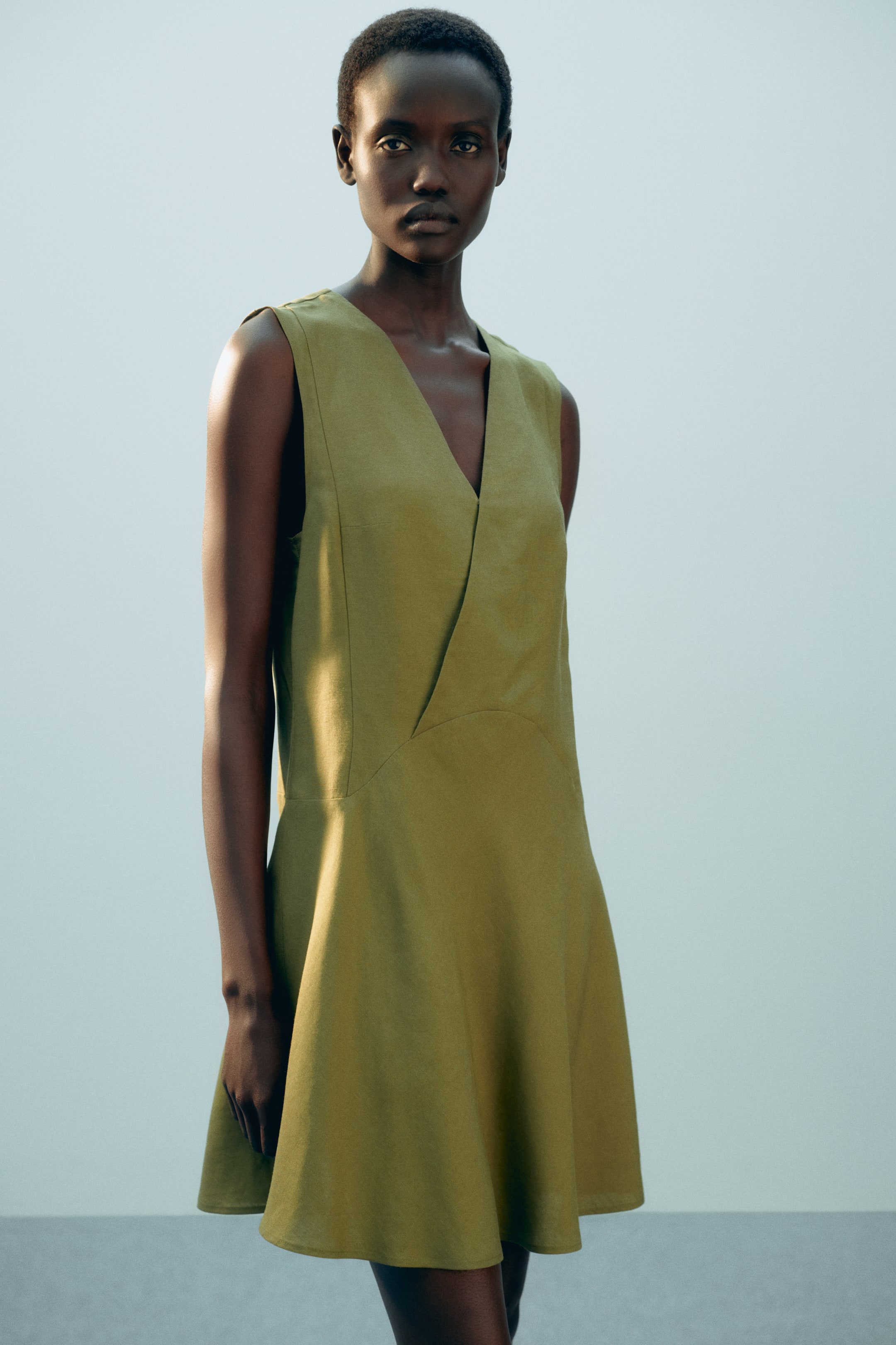 Xem ảnh lớn hơn: Người mẫu đang mặc một chiếc váy ngắn màu xanh ô liu, không tay. Chiếc váy này có kiểu dáng rộng rãi với cổ chữ V, phần trước được may chéo và các đường may tạo dáng phía trước, với phần chân váy nhẹ nhàng xòe ra.