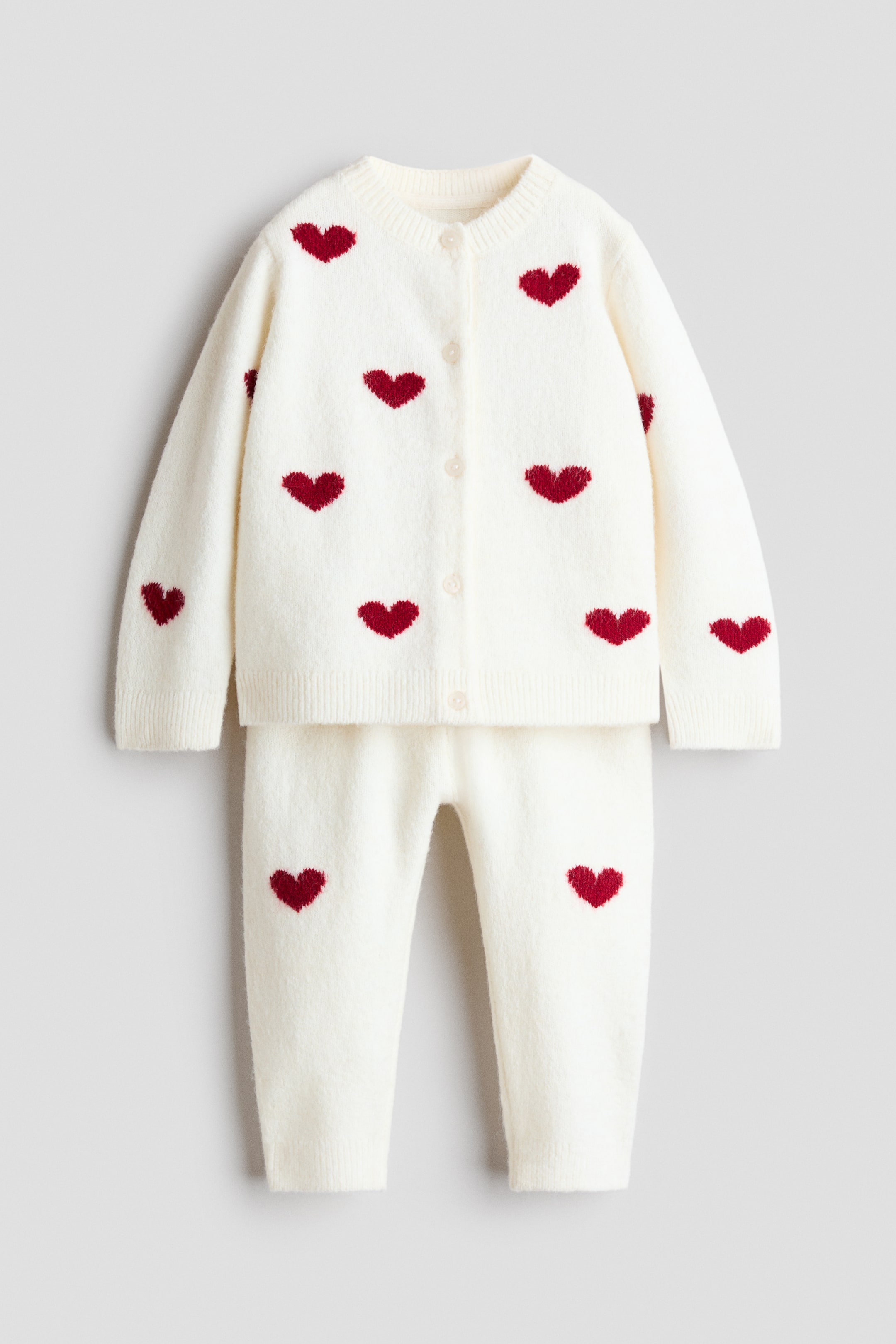 View larger image: A two-piece knit set is shown from the front, featuring a long-sleeve cardigan and matching trousers in a soft cream jacquard knit with red heart patterning. The cardigan features a round neckline, five front buttons, and ribbed cuffs and hem, while the trousers have matching ribbed ankle cuffs.