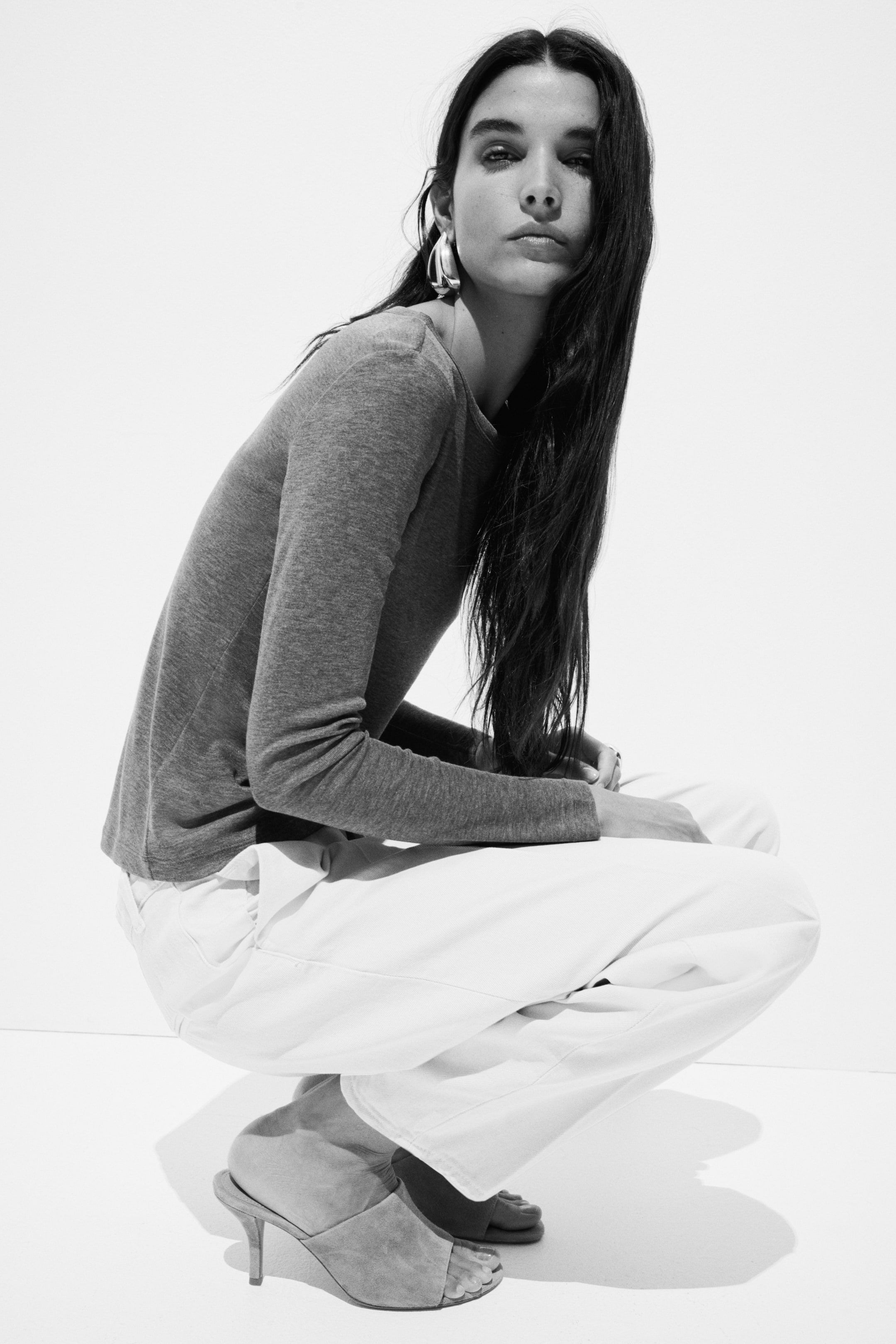 View larger image: A grey long-sleeved round-neck top, featuring a relaxed fit, is being worn. It is paired with white wide-leg pants that are pooling slightly at the ankles, and light-coloured open-toe mules with a slender heel are visible on the feet.