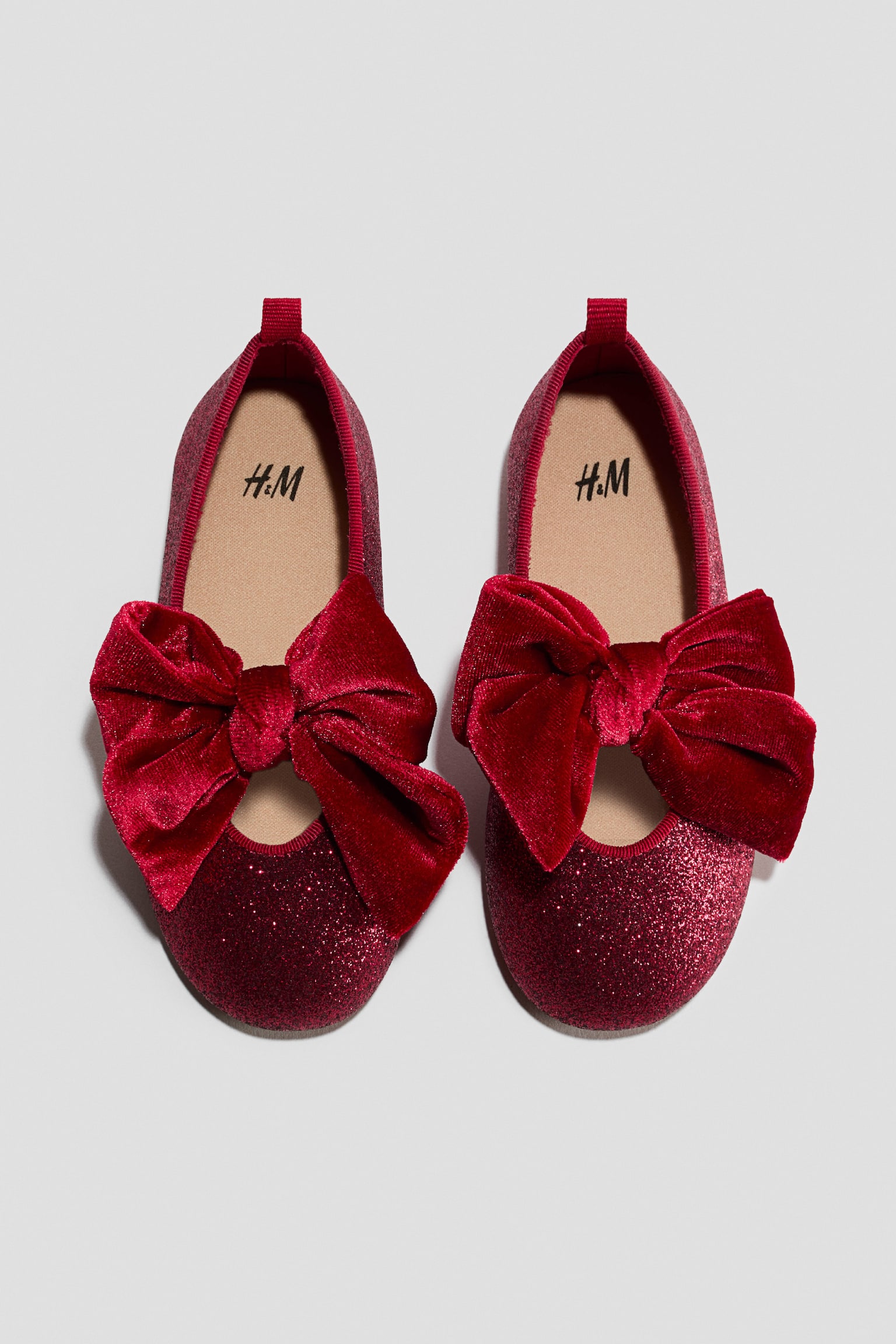 A pair of red glittery ballet flats is facing front, showcasing a large, plush red velvet bow draped across the top. The shoes display a tan interior lining with a black logo and a small ribbon pull tab at the back heel.