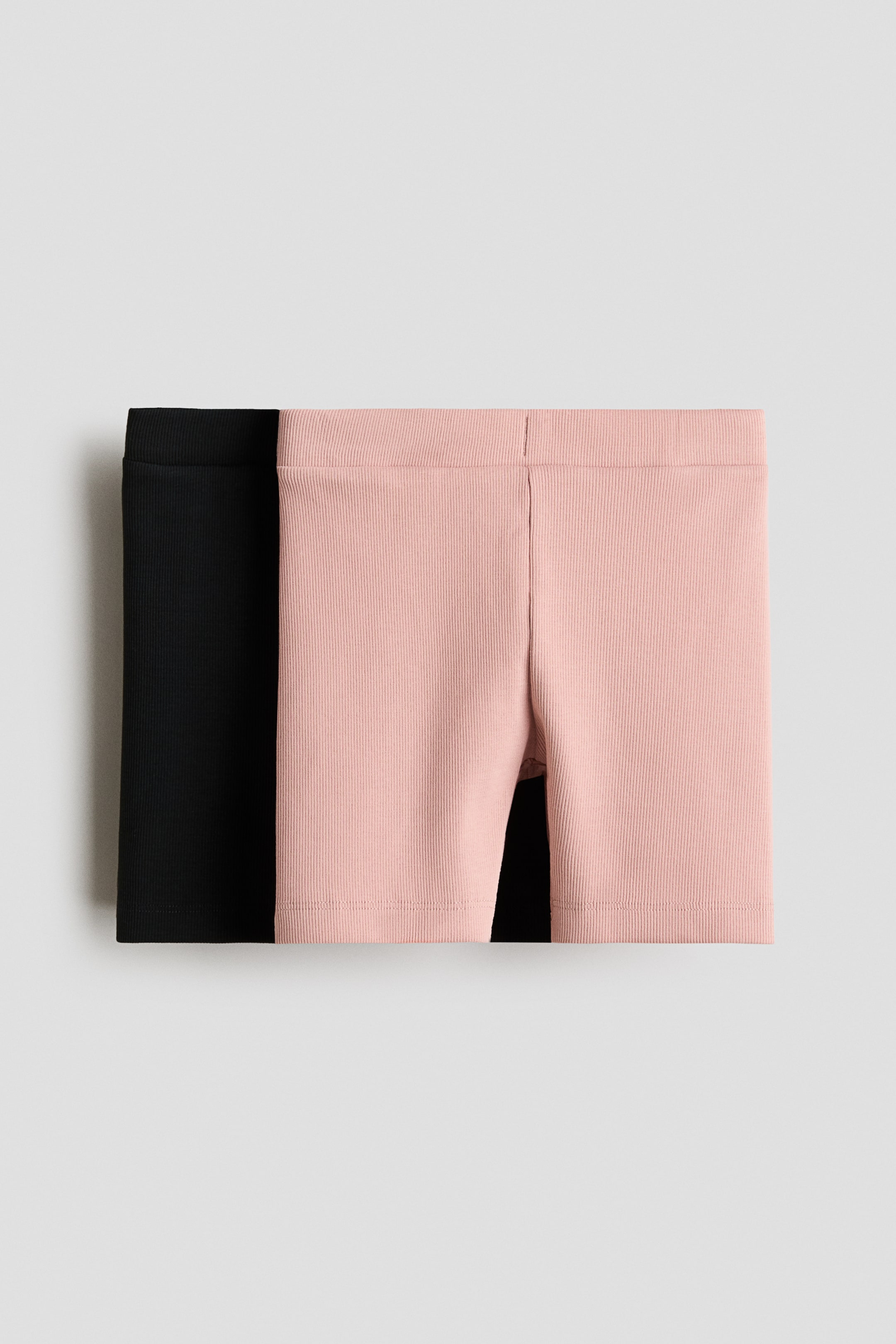 Visa större bild: Två par ribbade shorts ligger platt och framåtvända. Ett ljusrosa par överlappar delvis ett svart par. Det rosa paret visar en synlig centralsöm framtill, och båda paren har resår i midjan och en vertikal ribbad struktur.