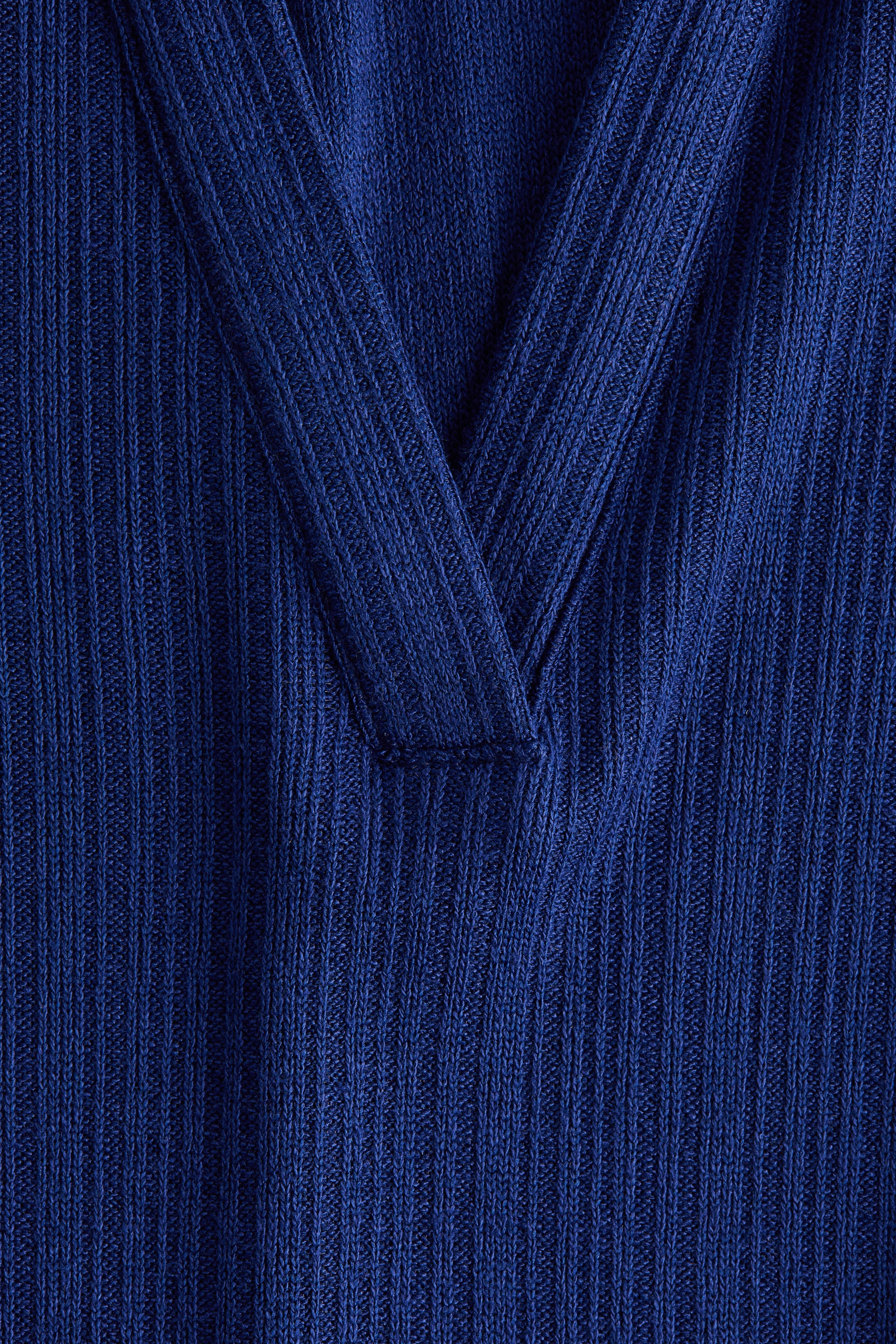 拡大画像を表示: 深い青色のリブ編みの衣服が、そのVネックラインの正面を見せています。ネックラインは、リブ編みの生地の2つの角度のある部分がVの基部で水平の縫い目で合わさって形成されています。