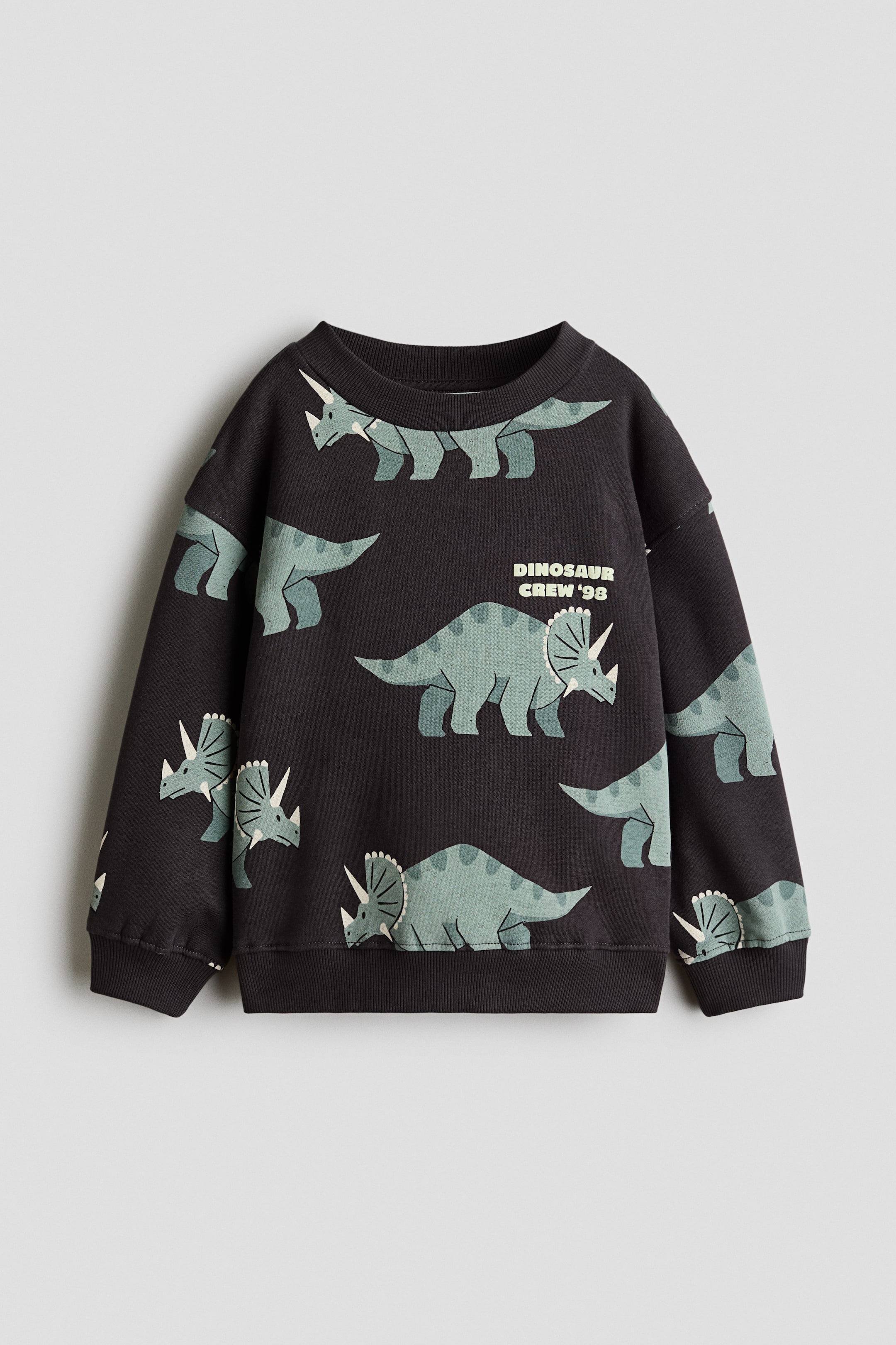 Büyük resmi görüntüle: Koyu gri bisiklet yaka bir sweatshirt düz bir şekilde sergileniyor; üzerinde açık yeşil stilize Triceratops dinozorlarından oluşan tüm yüzey baskısı bulunuyor. Sweatshirtte nervürlü yaka, nervürlü manşetler ve nervürlü etek ucu yer alıyor; ön kısmında 'DINOSAUR CREW '98' yazısı basılıdır.