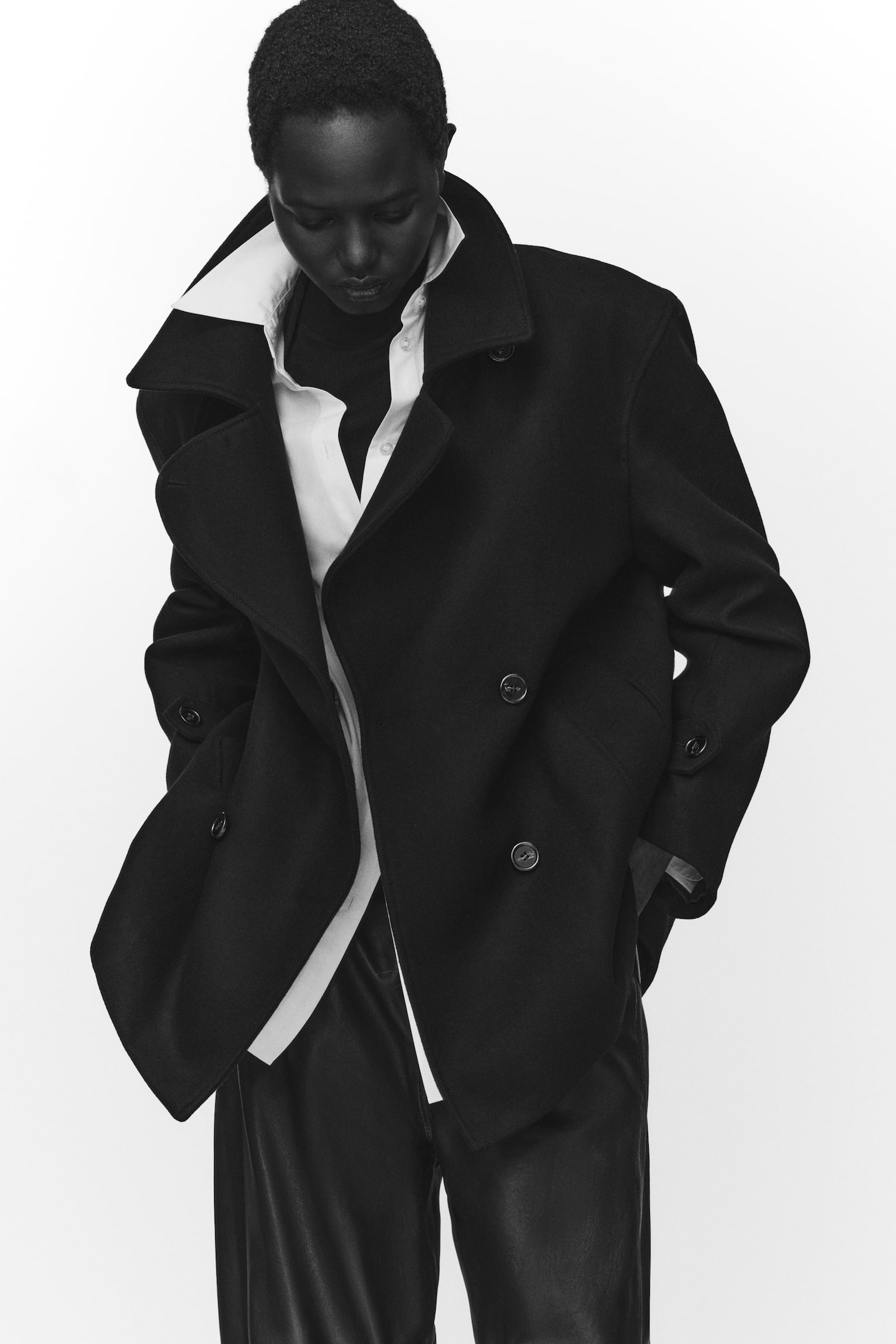 The model is wearing a dark wool-blend double-breasted coat unbuttoned, featuring dropped shoulders, large front buttons, and buttoned tabs at the cuffs. The coat is layered over a white collared shirt and a dark turtleneck, and paired with dark trousers. - 1