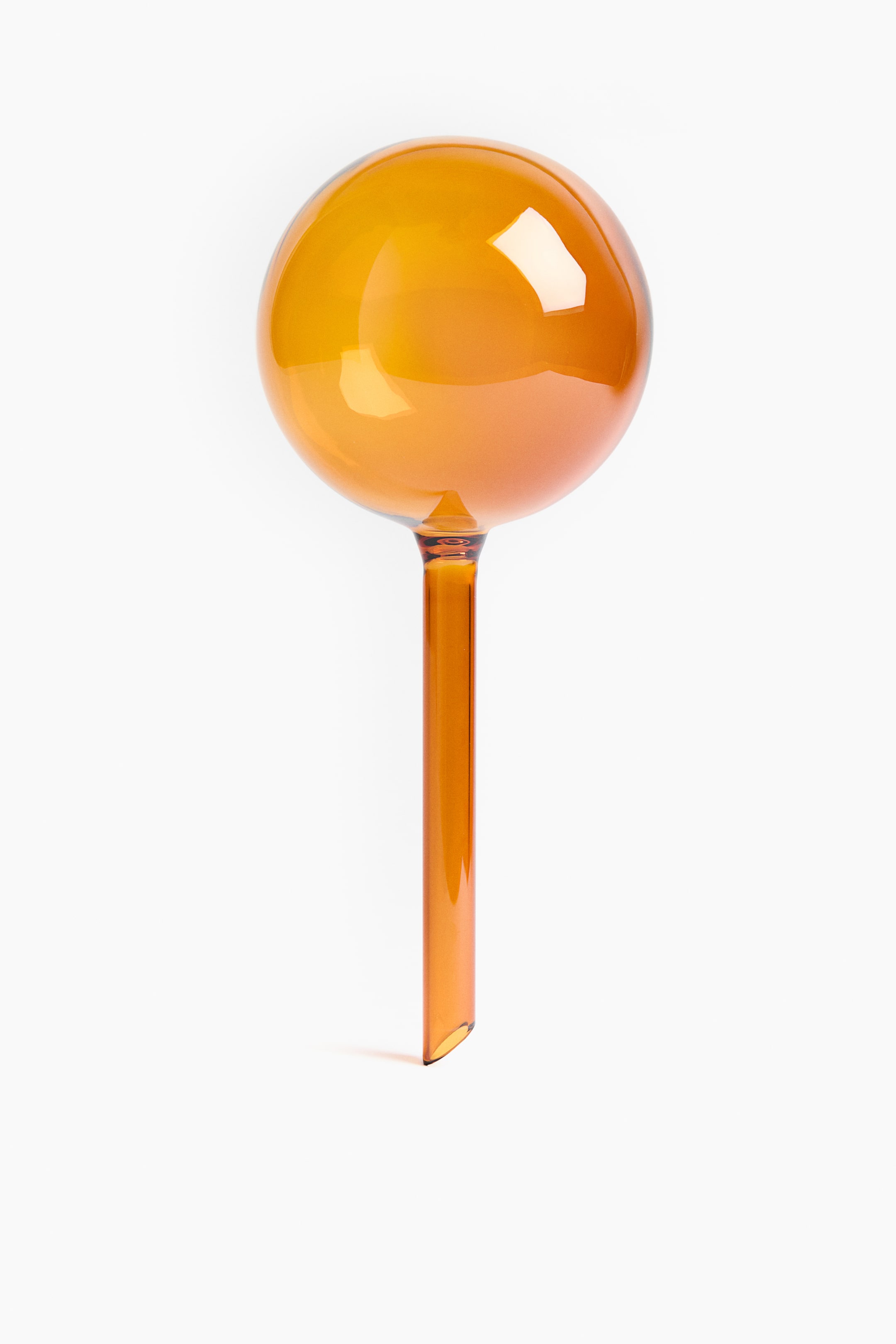 Agrandir l'image: Un globe d'arrosage en verre ambré présente un réservoir sphérique au sommet. Une tige droite et étroite s'étend vers le bas depuis la sphère et présente une coupe diagonale à sa base.