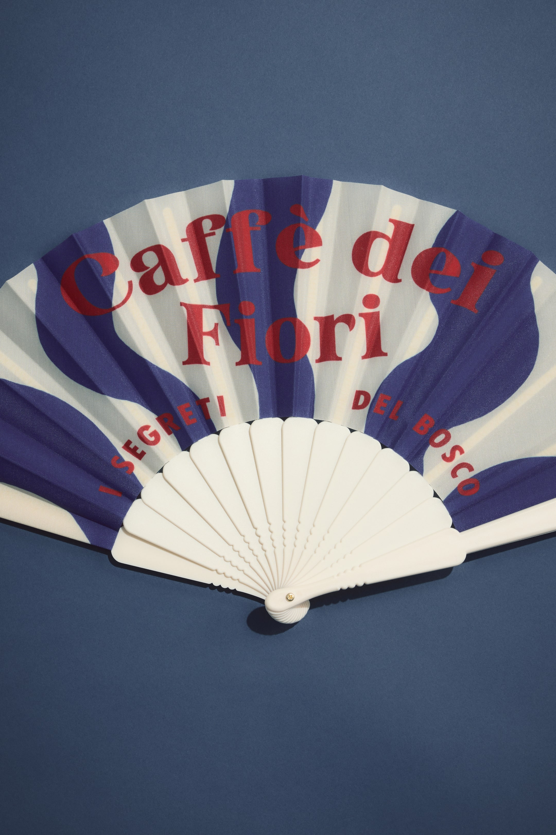 Agrandir l'image: Un éventail à main est partiellement ouvert, incliné vers le spectateur, sa feuille blanche affichant des rayures bleues ondulées et un texte rouge indiquant 'Caffé dei Fiori'. Les brins de garde en plastique blanc forment la base, reliés par une goupille pivotante de couleur dorée.