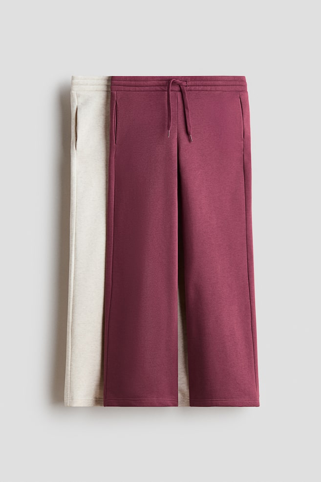 Two pairs of straight-leg track pants are hanging flat, with a maroon pair layered in front of a light beige pair. The front-facing maroon pair is showing an elasticated waistband with a matching drawstring, straight legs, and discreet side pockets. - 1