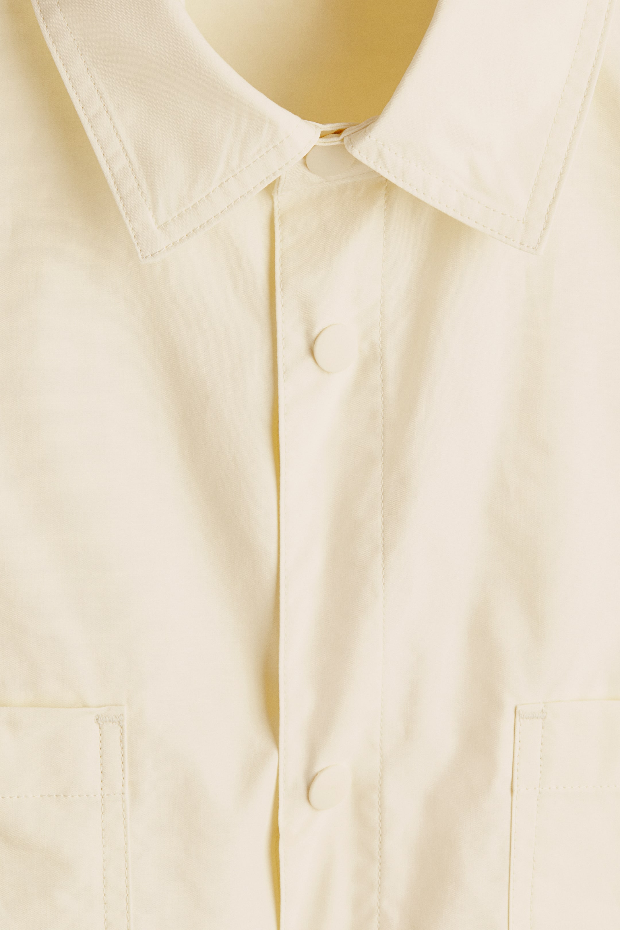 Xem ảnh lớn hơn: Một chiếc áo sơ mi màu vàng nhạt, mặt trước đang hướng về phía trước, có cổ áo nhọn với đường chỉ viền. Phần xẻ giữa được cài bằng hai nút tròn cùng màu hiện rõ, và hai túi vuông vá được hiện rõ ở phần dưới.