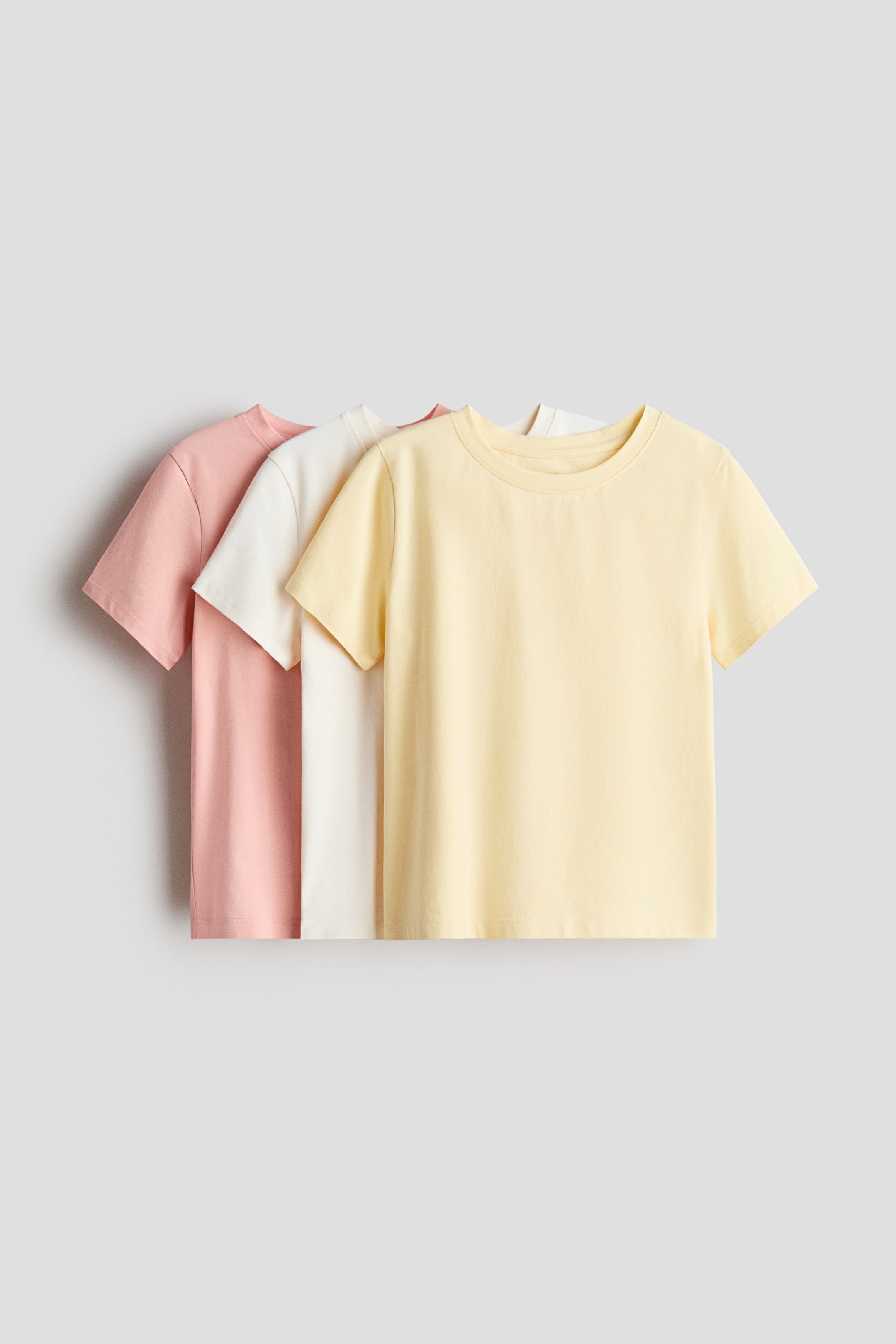 View larger image: Three short-sleeved cotton jersey t-shirts are layered and facing front, showing a light yellow tee in the foreground, a white tee in the middle, and a pink tee in the back. Each t-shirt is designed with a round neckline featuring a narrow trim.