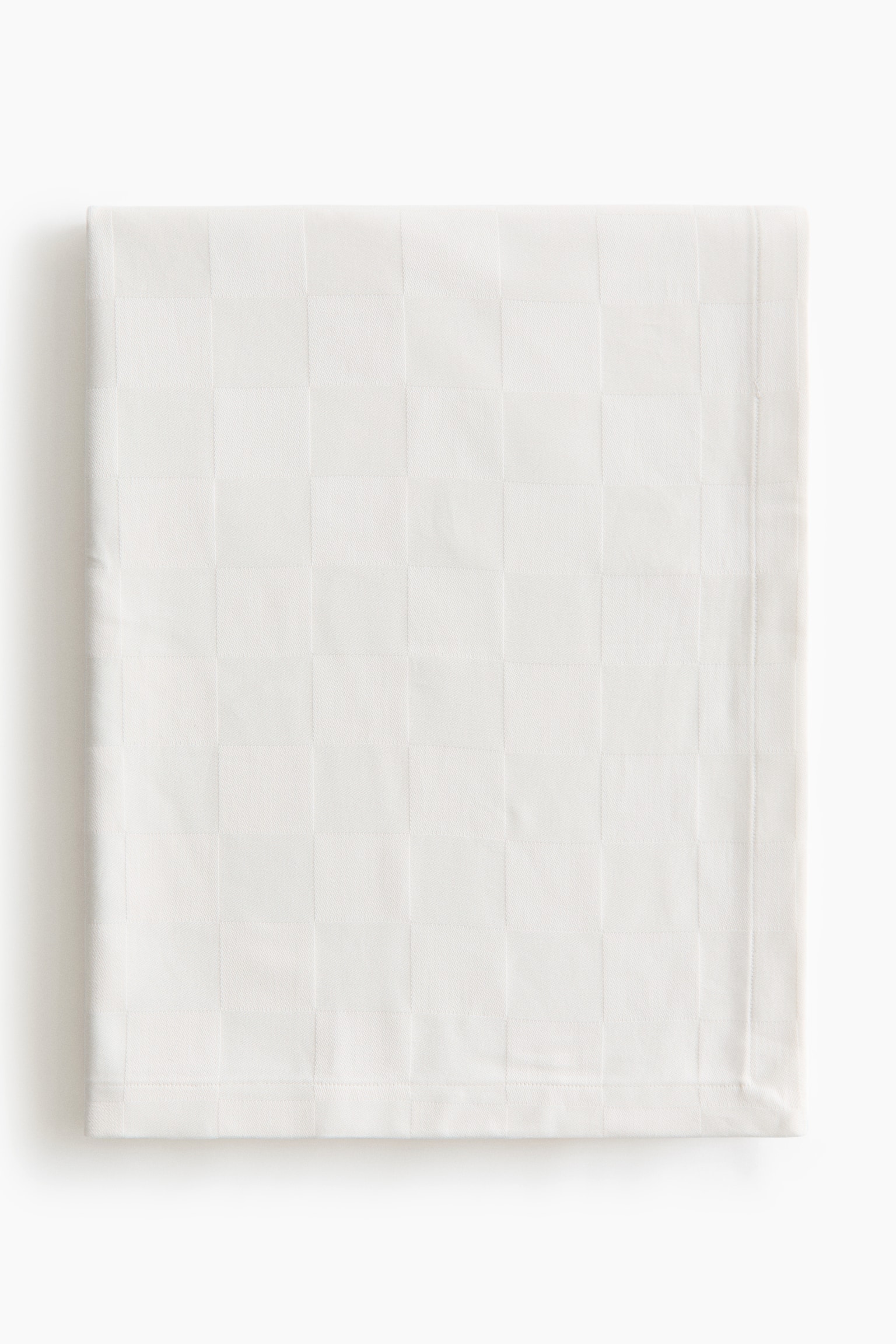 A white cotton tablecloth is lying folded flat and facing front, featuring a subtle tone-on-tone woven check pattern. The tablecloth is displaying a stitched, folded hem along its visible bottom and right edges.