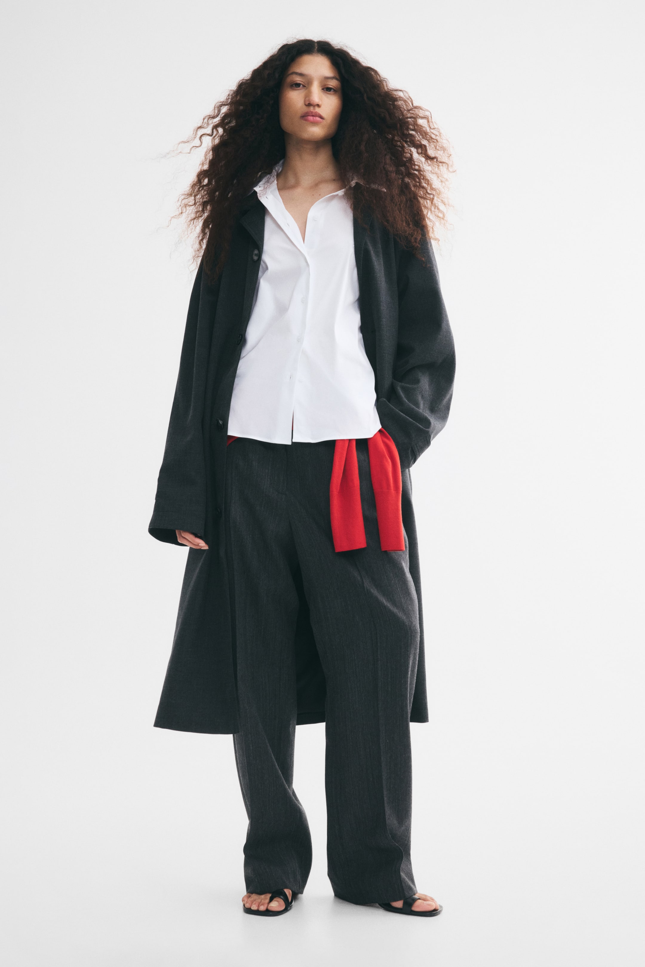 View larger image: A white long-sleeve shirt with a collar and button-down front is worn by the model. The shirt is layered under an open dark grey coat and paired with dark grey trousers and a red knit tied at the waist.