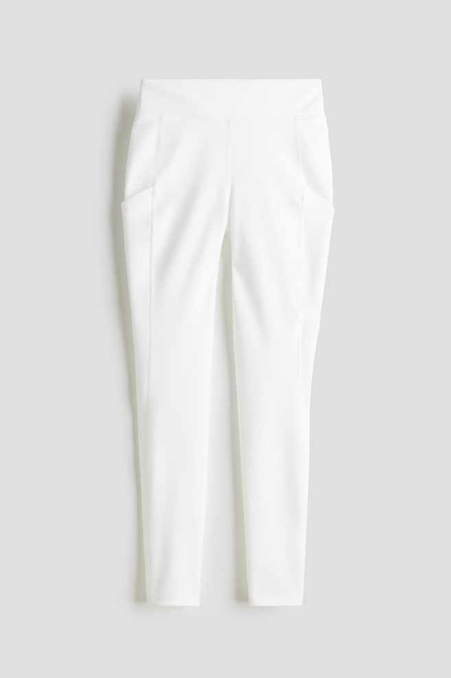 Weisse Reitleggings werden vorne präsentiert, mit hohem Bund und breitem Taillenbund sowie Seitentaschen an beiden Oberschenkeln. Hergestellt aus einem glatten Funktionsstoff, sind die Innenseiten der Beine und der Sitzbereich sichtbar mit einem vollständigen Silikongriffmuster texturiert, und die Säume sind mit elastischem Band abgeschlossen. - 1