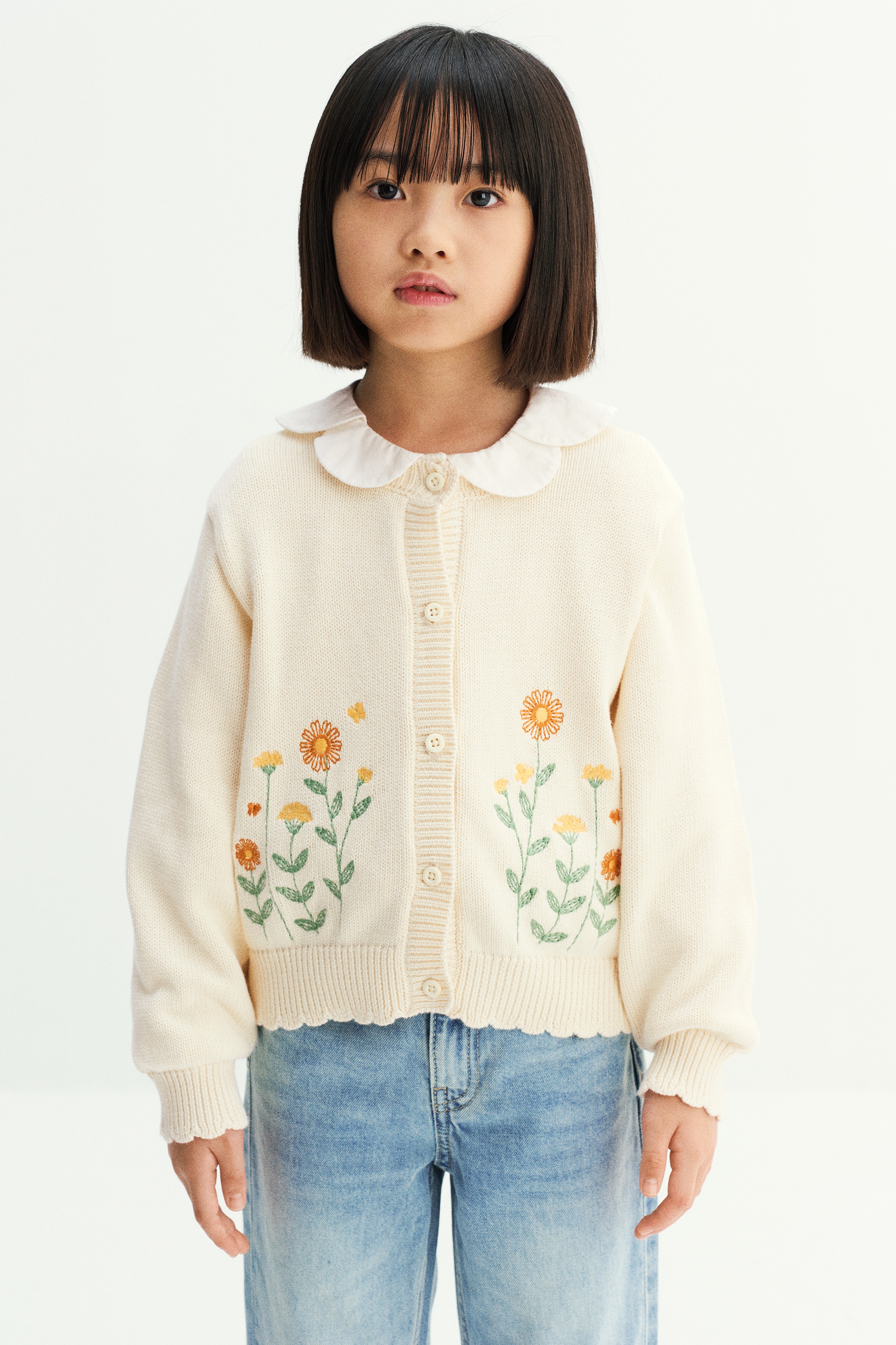 A cream-colored cotton knit cardigan is shown from the front, featuring embroidered orange and yellow floral motifs on the lower front. It has a round neckline, a button-down front, dropped shoulders, and ribbed trims with scalloped edges at the neckline, cuffs, and hem.